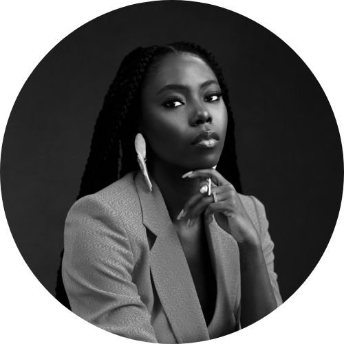 Black woman with braided hair wearing a blazer and large earring, looking contemplatively at the camera in a studio portrait.