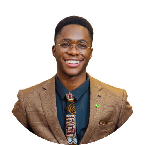 A smiling young man wearing glasses, a brown plaid suit, a dark blue shirt, and a colorful patterned tie. He has a small flag pin on his lapel and is standing against a white background with a black curved detail in the top left corner.