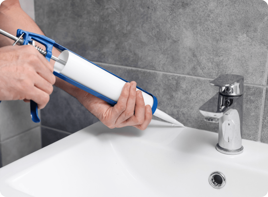 Persoon gebruikt een kitspuit op een wastafel in een badkamer met grijze tegels.