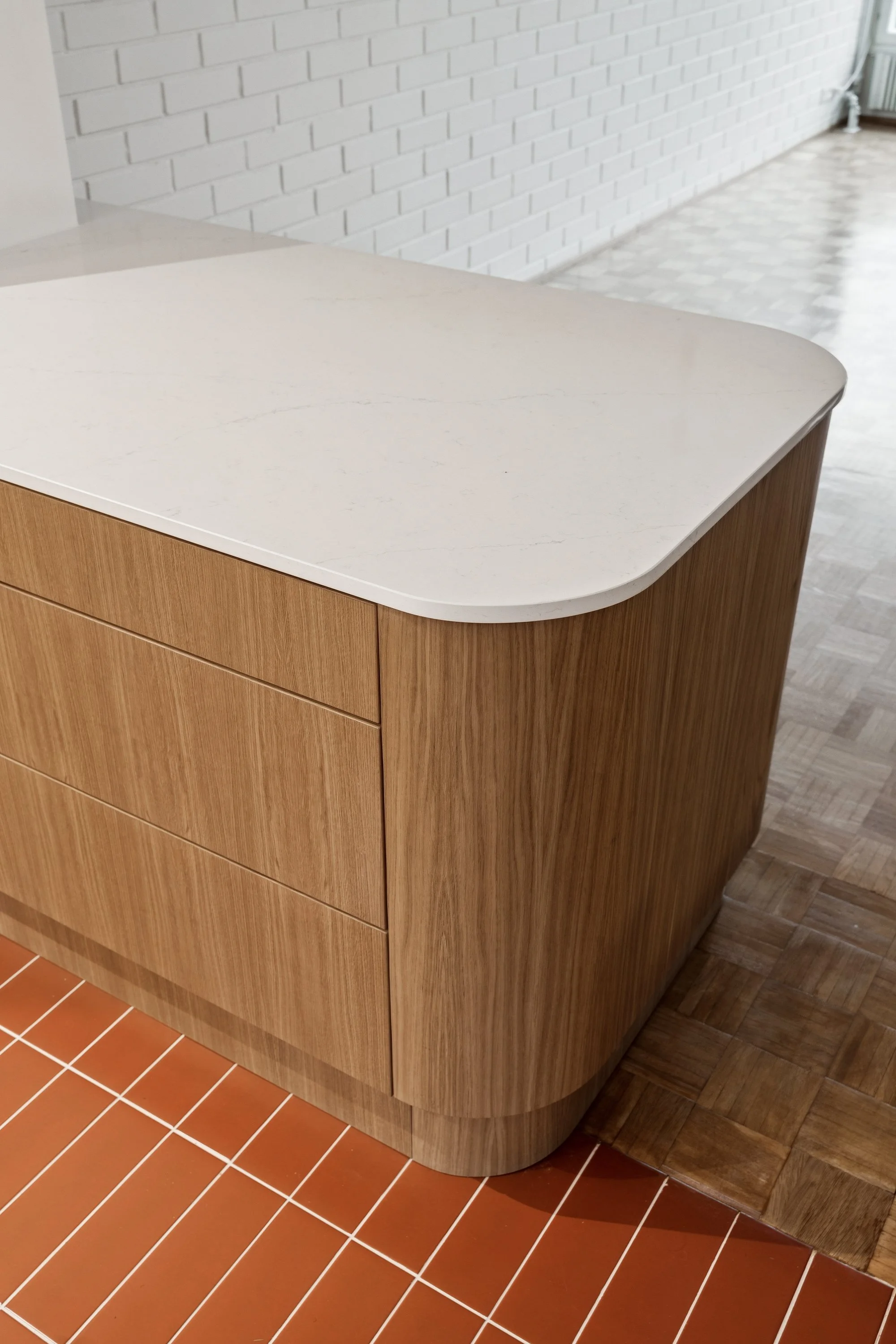 Kuvassa on pyöristetty keittiön niemeke, jossa on valkoinen Silestone Persian White ja tammiviilusta tehdyt ovet. Taustalla on valkoinen tiiliseinä ja ikkunasta näkyy osa huonetta. Punainen terracottalattia