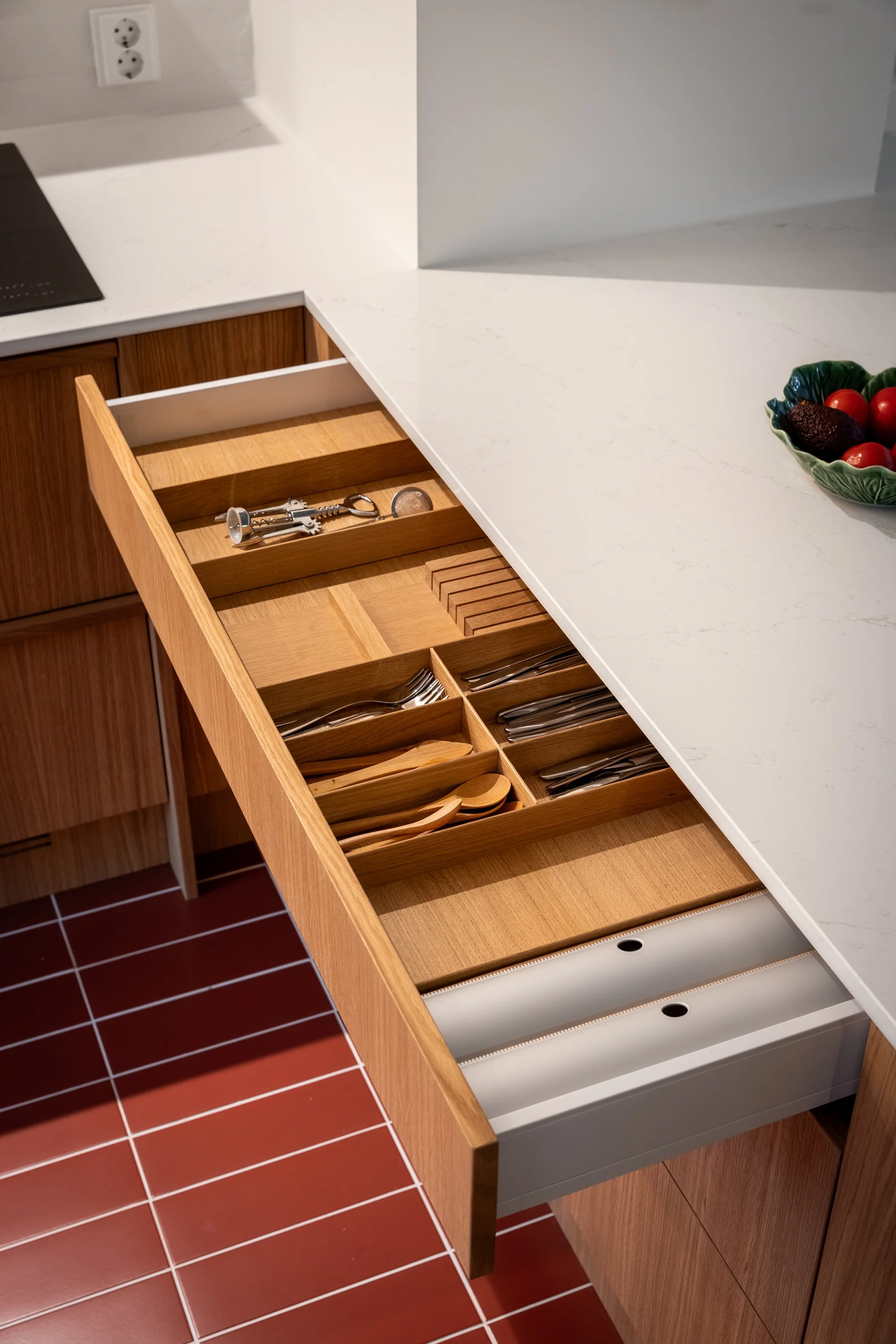 Avautuva keittiön puinen vetolaatikko sisältäen haarukat, veitsen ja lusikat, vaalea puu ja valkoinen Silestone Persian White työtaso, punainen terracottalaattalattia.