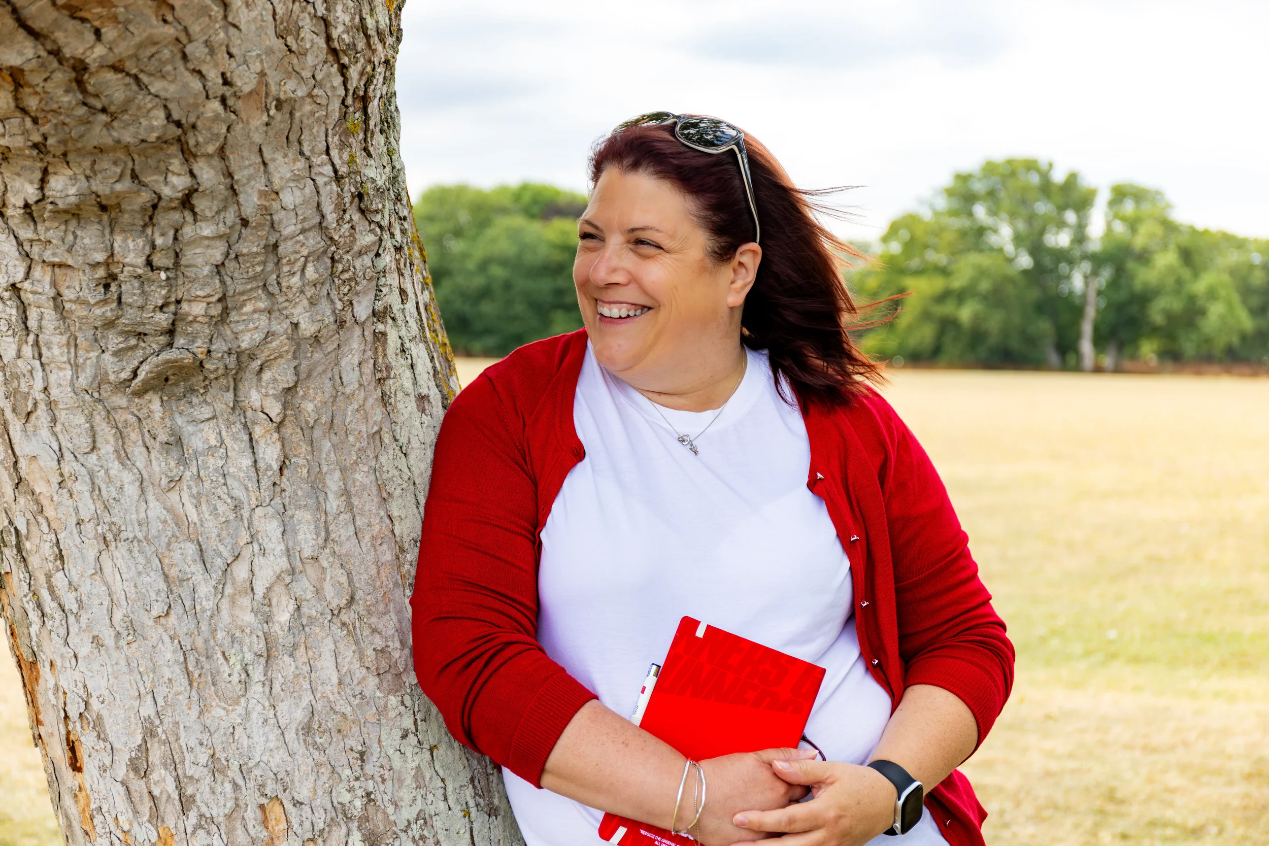 A woman with reddish-brown hair and sunglasses on her head is smiling while leaning against a tree. She is wearing a white T-shirt and a red cardigan, holding a red notebook and a white pen, with a scenic background of green trees and brown grass.