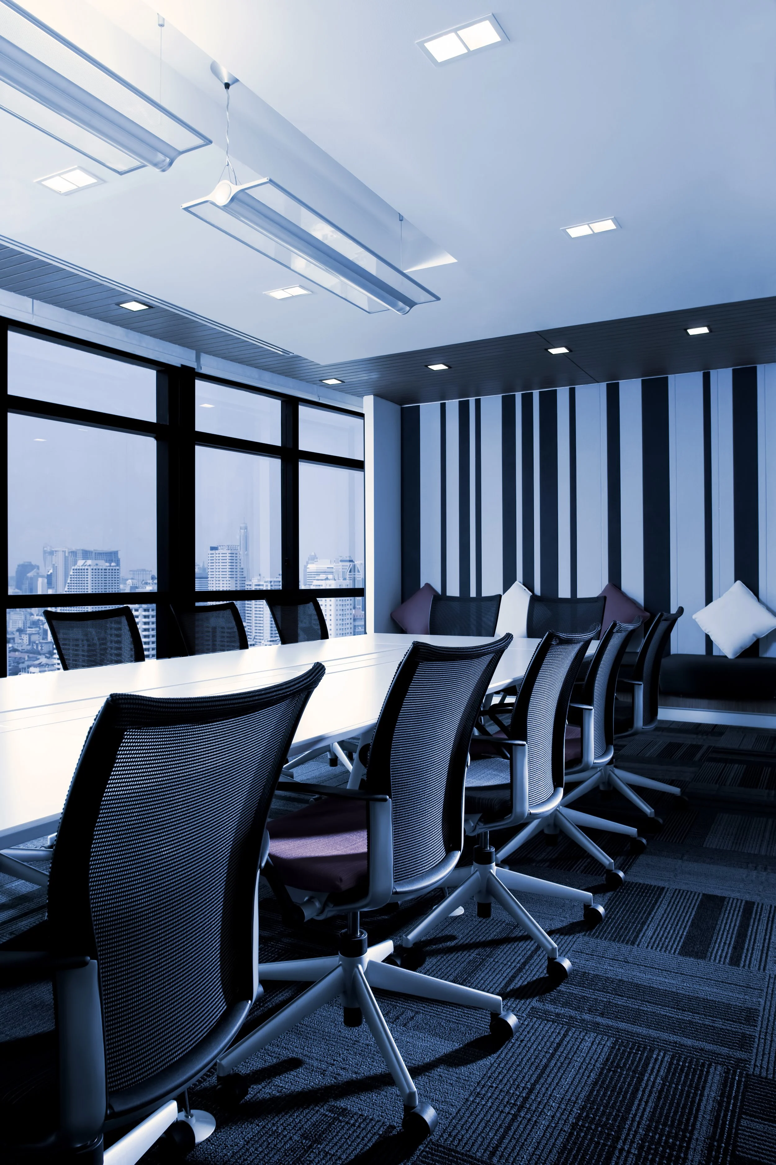 Modern conference room with a long white table, black mesh chairs, large windows showing a cityscape, striped accent wall, and ceiling lights.