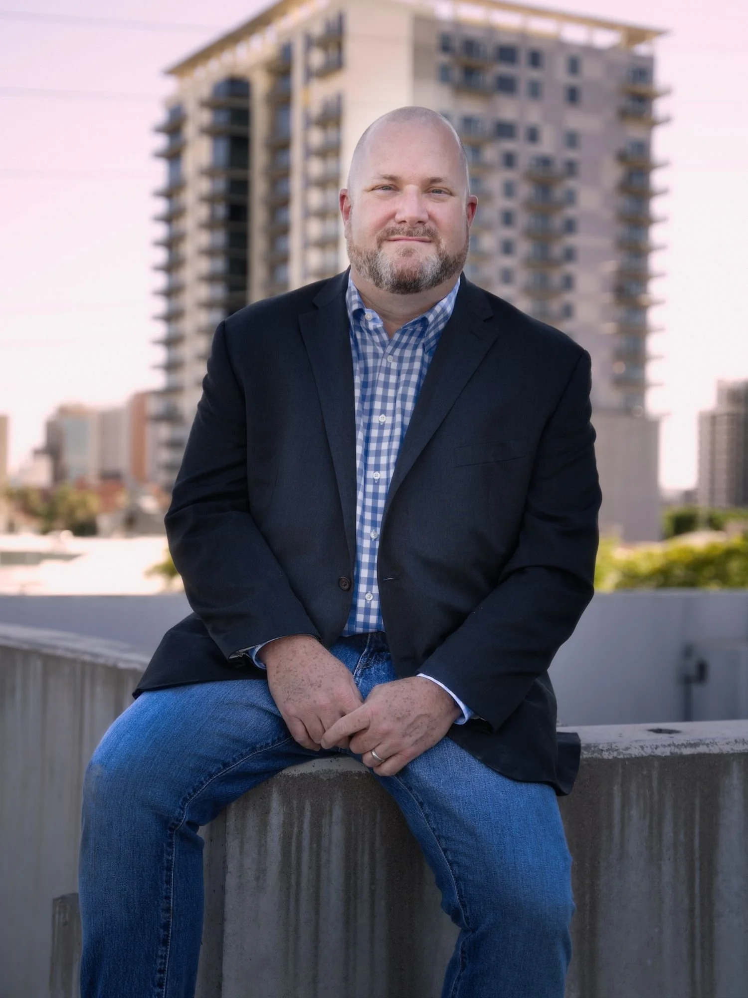 A man with a beard wearing a blue blazer over a blue checkered shirt, sitting on a concrete ledge with a cityscape background.