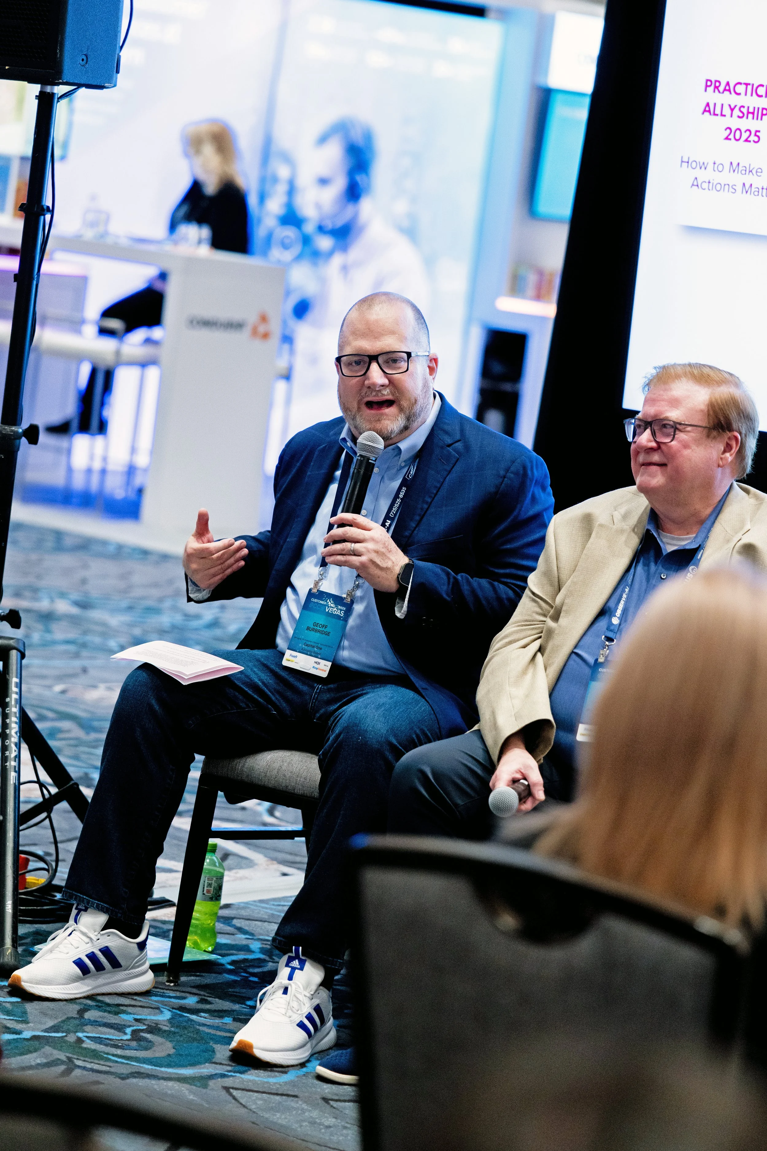 Man in glasses and dark suit speaking into microphone during panel discussion at conference, with other panelist seated next to him and audience in foreground.