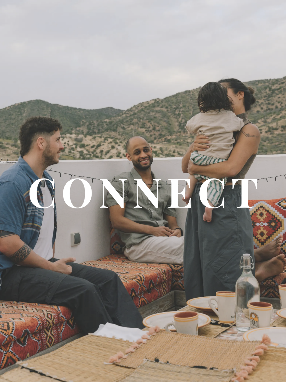 A group of four people gathered on an outdoor patio with a desert mountain backdrop. Two women and two men are engaged in conversation, with one woman holding a child. There are cups, a water bottle, and a table with a woven tablecloth in the foregro