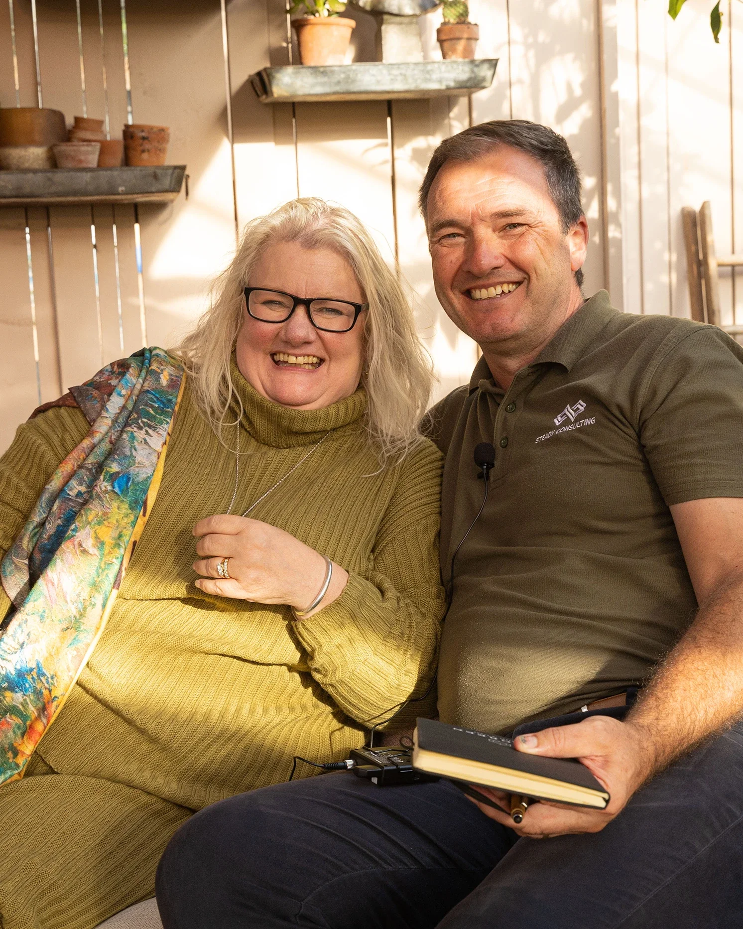 Photo credit: Tammy Marler. Executive and business Coach, mentor and consultant Neil Stead smiling and laughing with a woman (Kate Cooney - Corporate Partnerships Manager at Perennial) as they record a podcast in the sunlight within a garden shed.