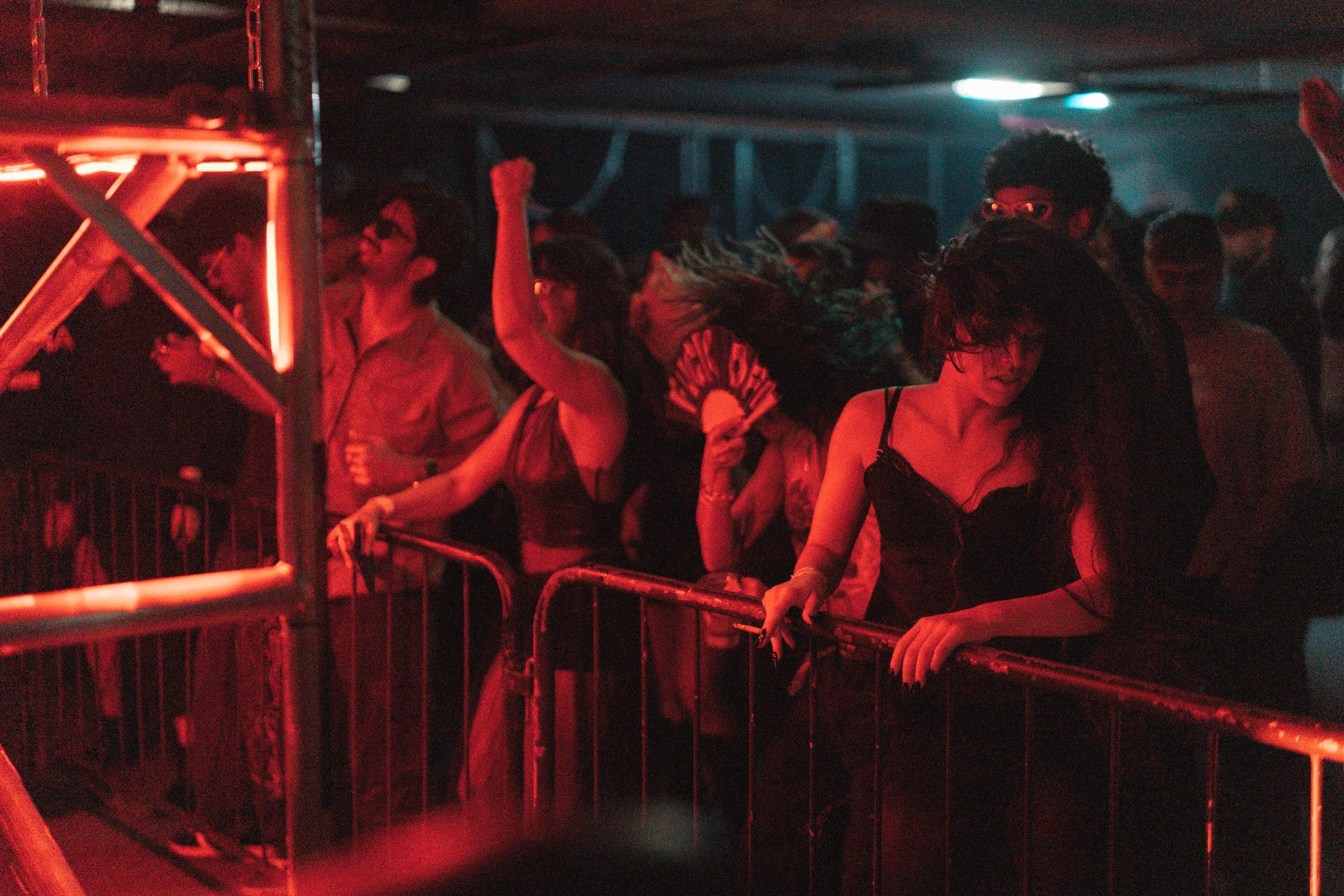 People dancing passionately at a nighttime event, with some holding a fan that says 'Easy Girl' and a woman with long hair in a black dress near a metal barricade.