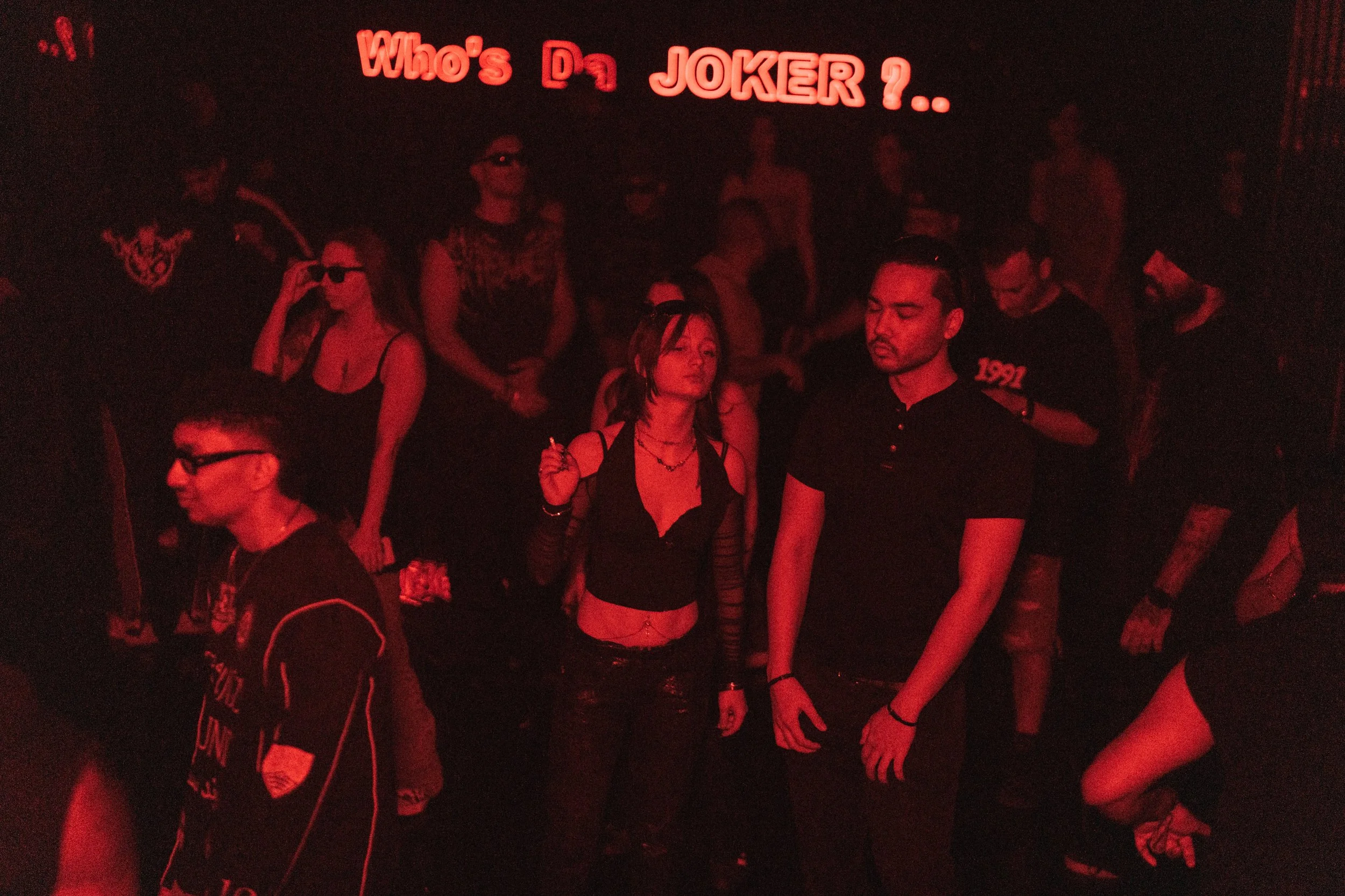 People at a nightclub illuminated by red lighting, with a neon sign reading 'Who's Da JOKER ?..' overhead.
