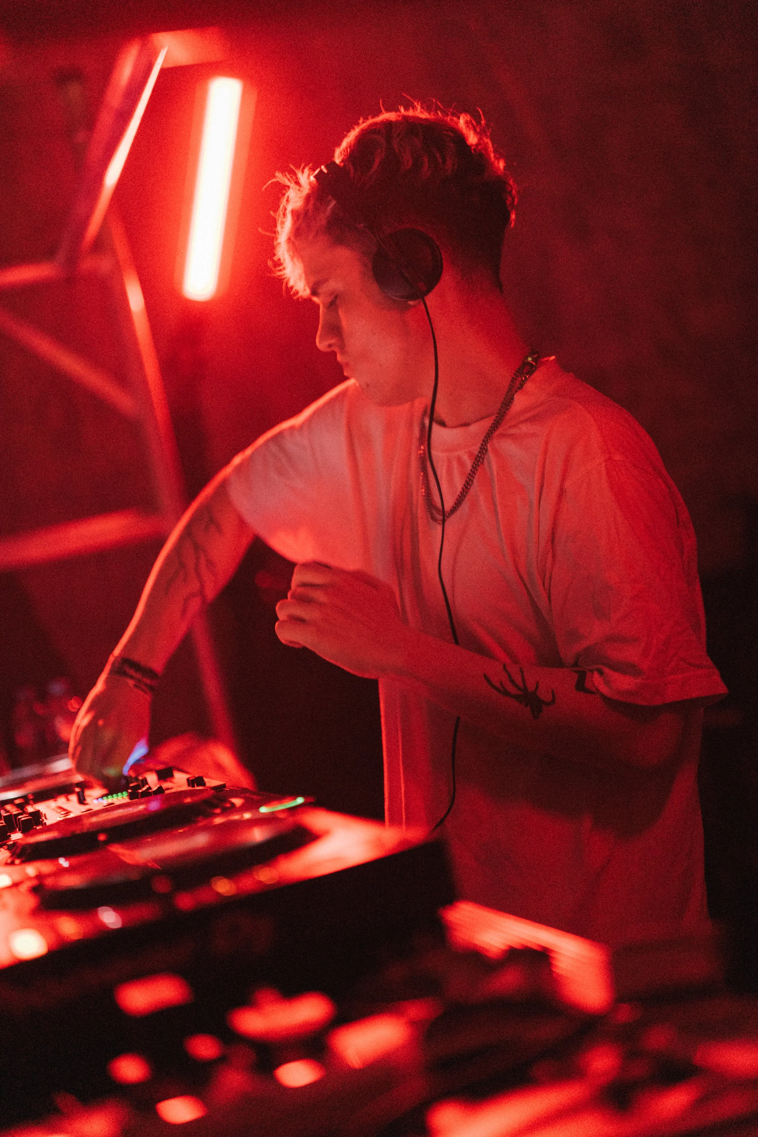 A young male DJ with tattoos on his arms, wearing a white t-shirt and headphones, is performing at a nightclub or music event bathed in red lighting, standing behind a DJ booth with turntables and audio equipment.
