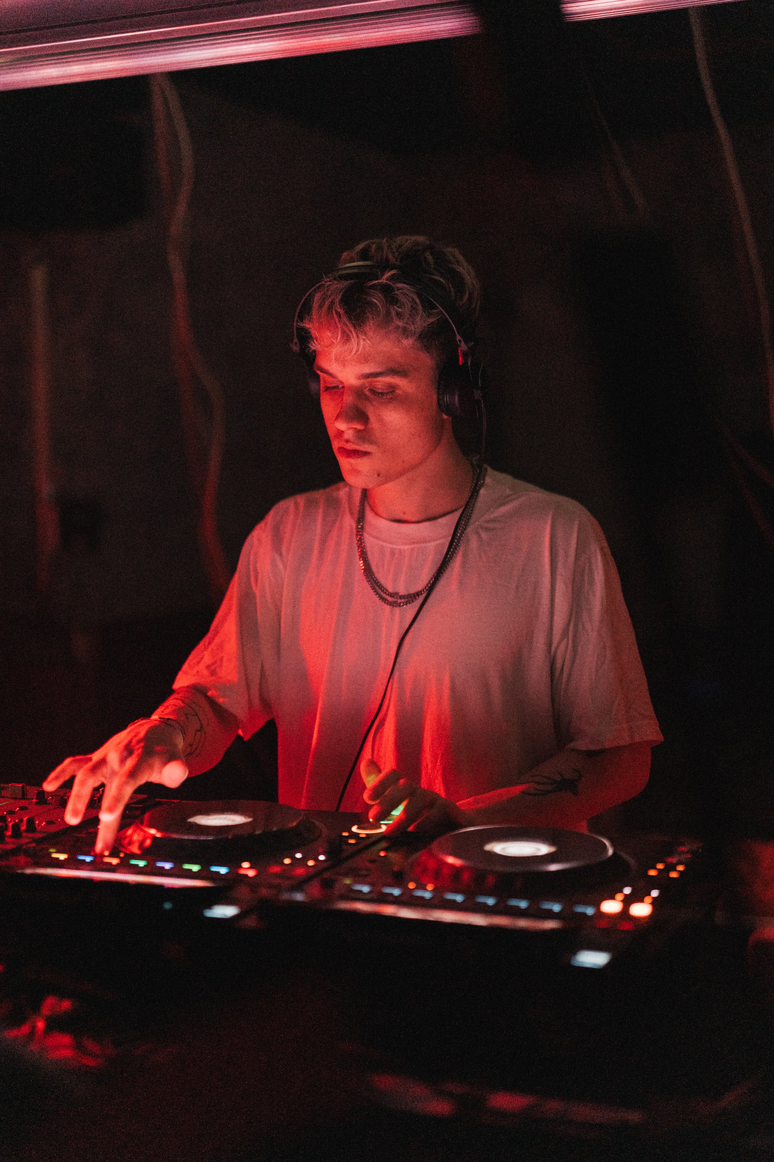 A young male DJ wearing headphones, a white t-shirt, and layered necklaces, mixes music on a DJ controller in a dark setting illuminated by red lighting.