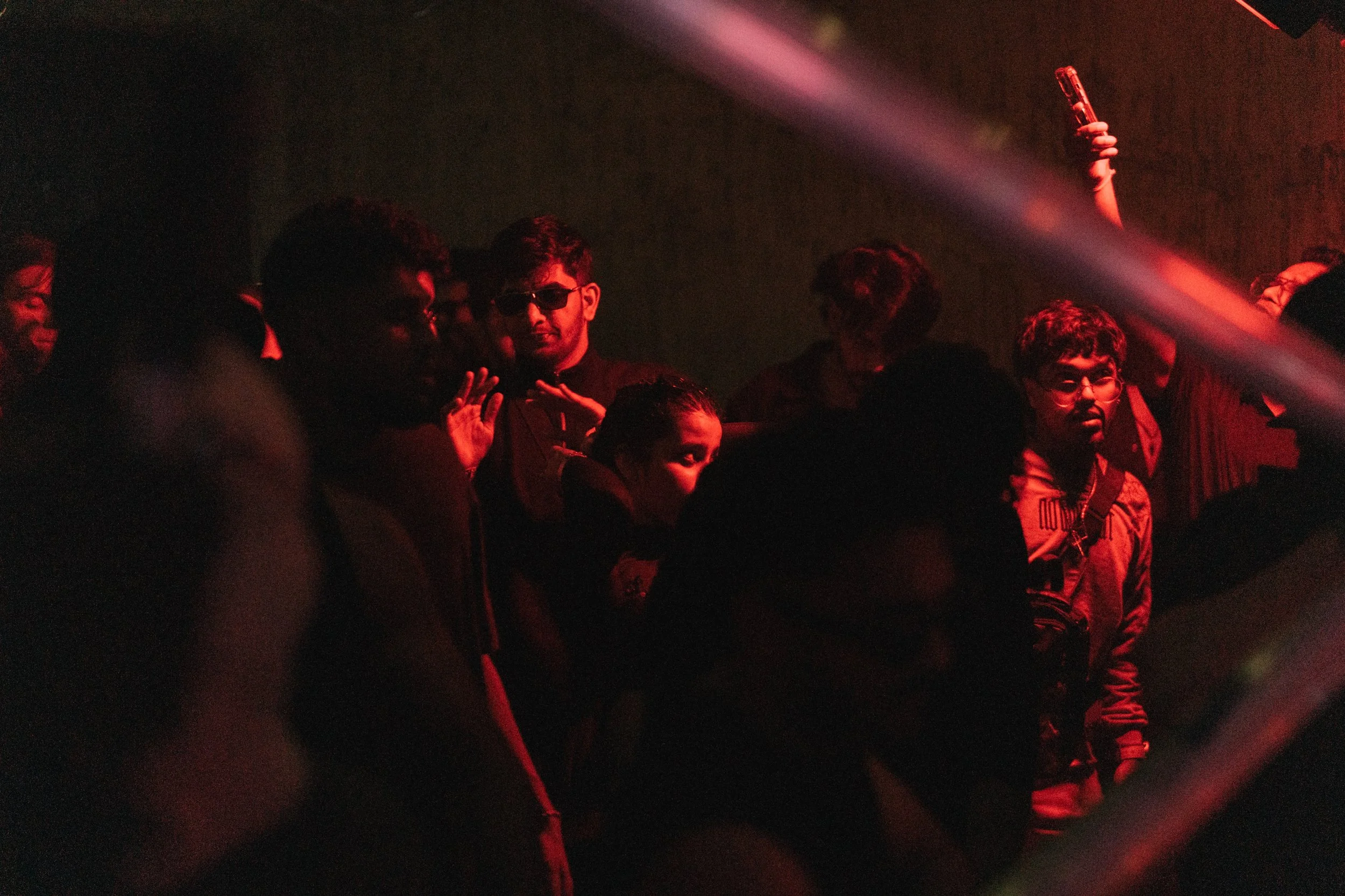People standing in a dark room illuminated by red light, some with sunglasses, some taking photos with phones.