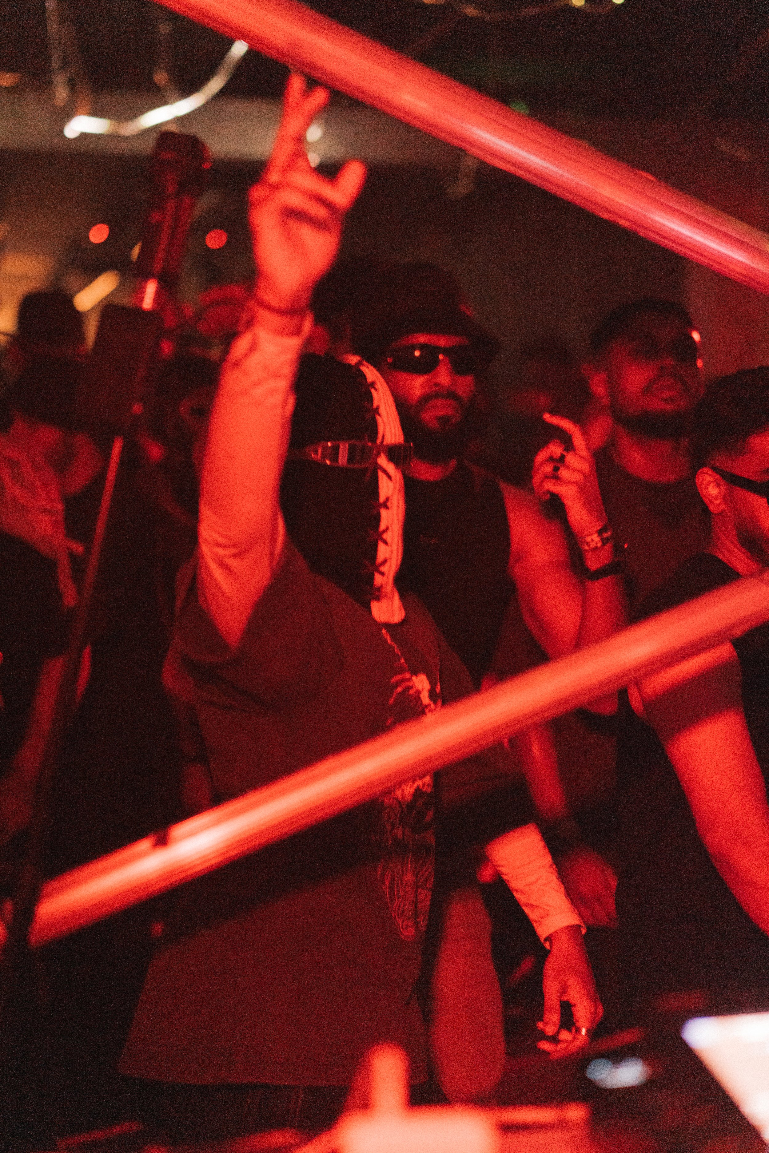 People dancing at a nightclub with red lighting, some wearing sunglasses and bandanas, surrounded by others in the dark.