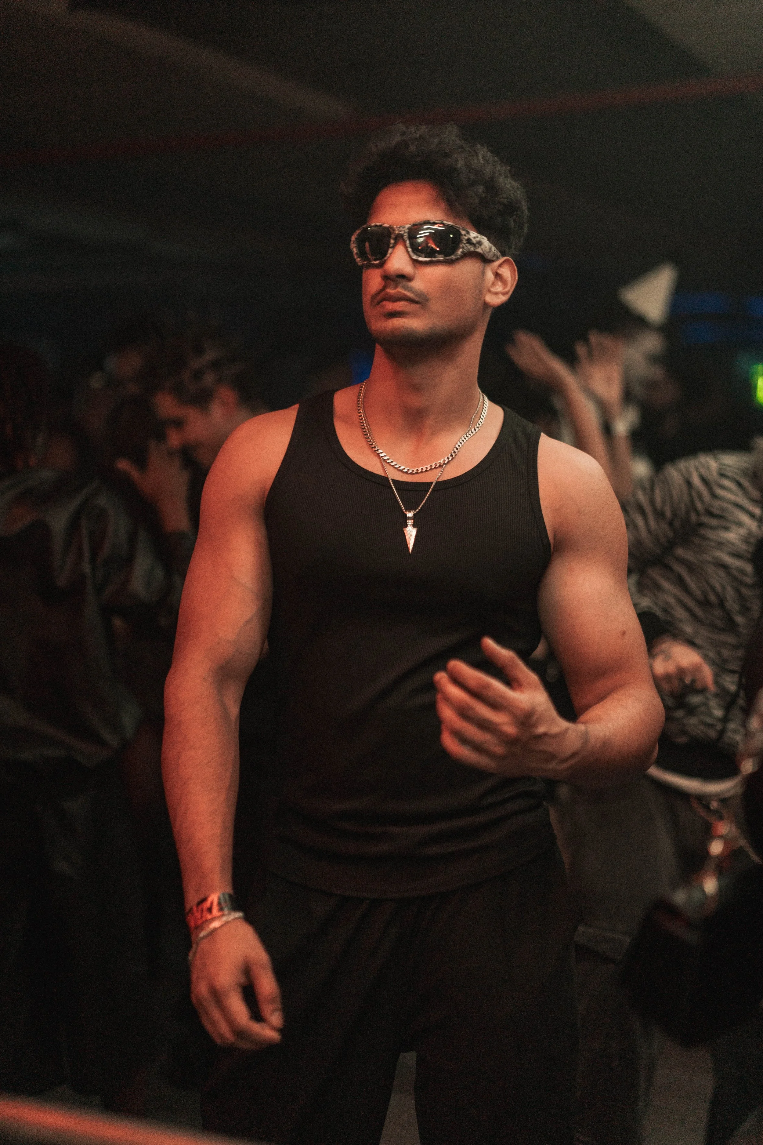 A young man wearing sunglasses, a black sleeveless shirt, and silver jewelry, standing in a dimly lit nightclub or party with other people in the background.