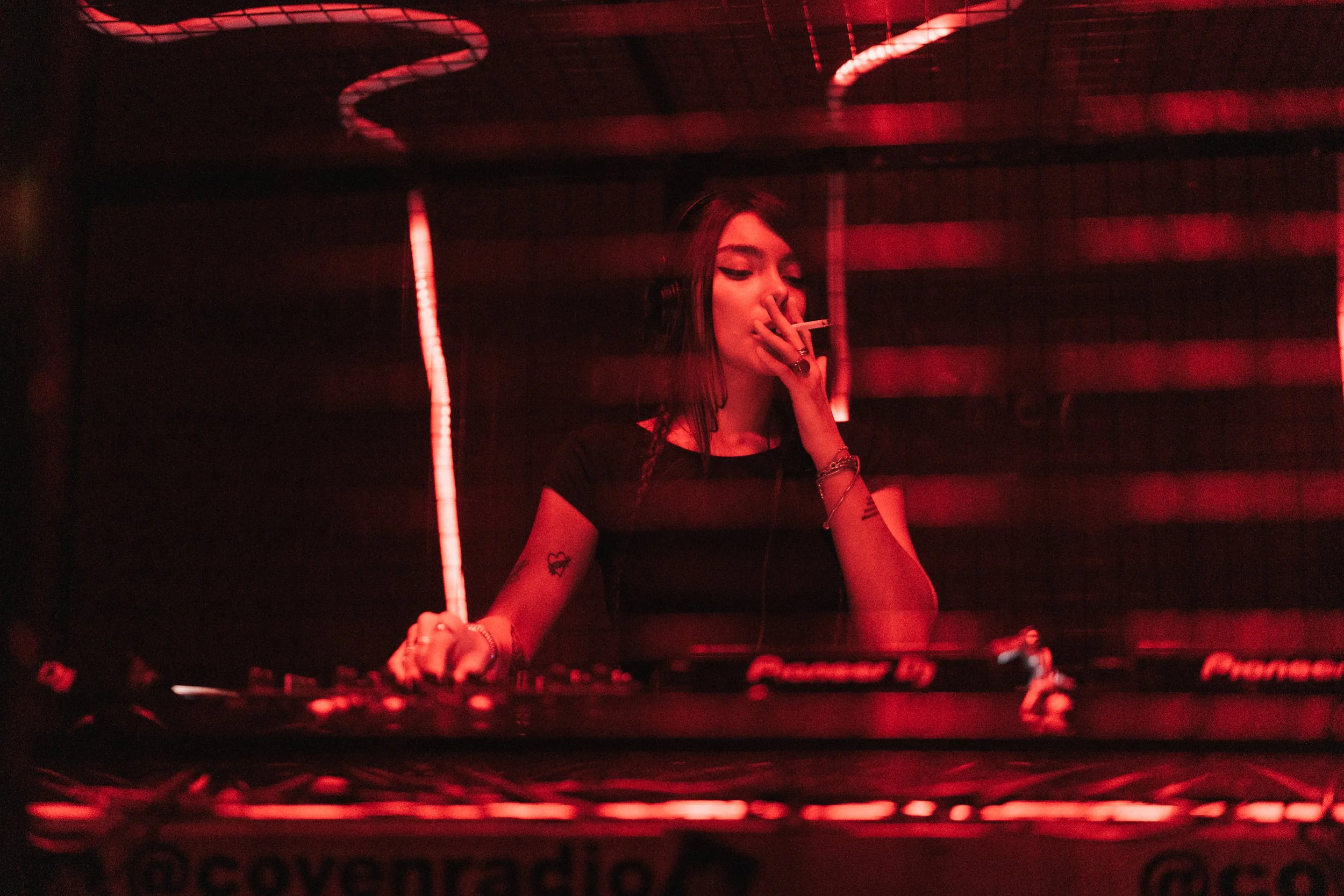 A female DJ with long dark hair and tattoos on her arms is playing music at a club, wearing headphones and smoking a cigarette, illuminated by red lighting.