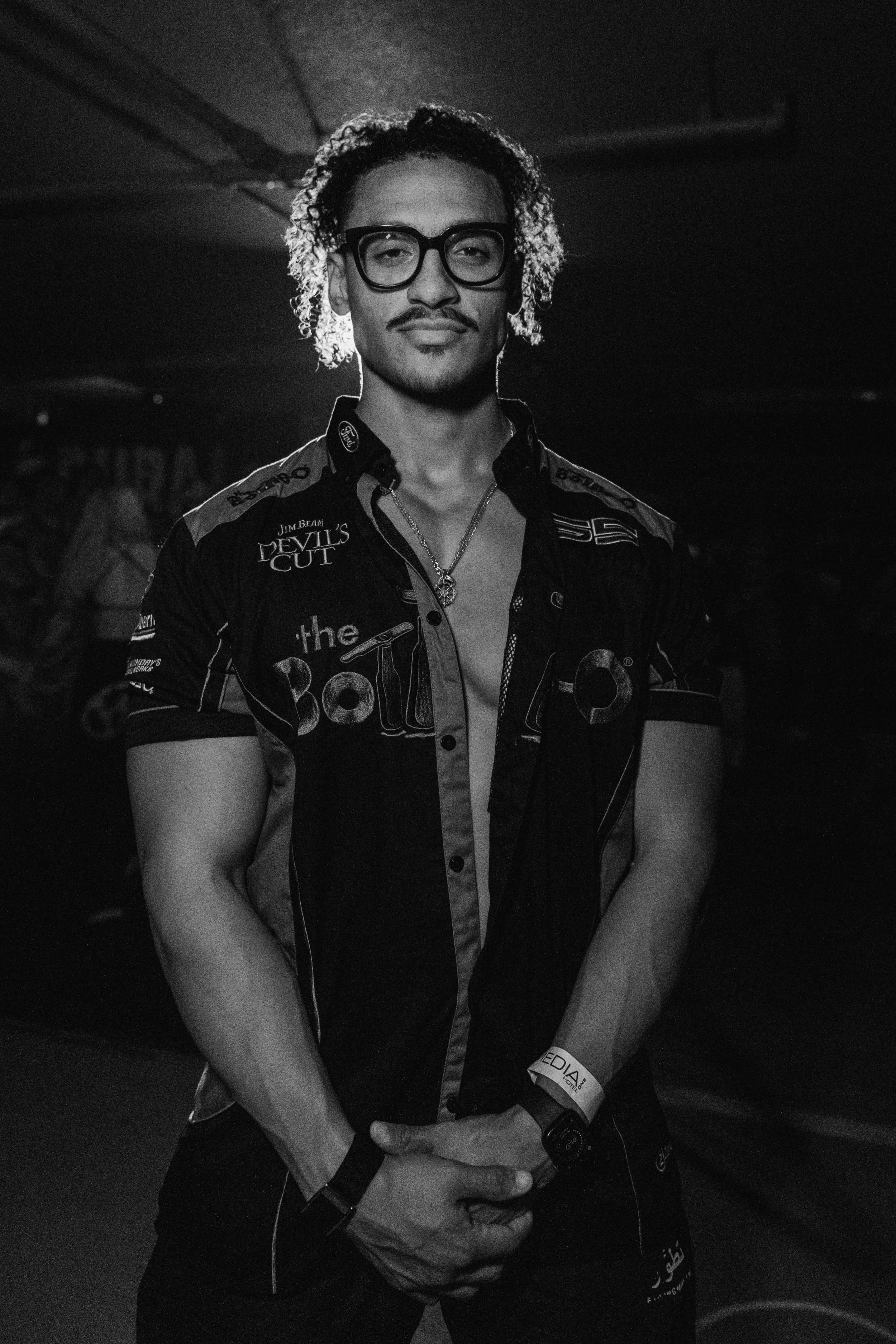 A man with glasses and curly hair stands in a dark room, wearing a unbuttoned racing shirt with various logos and a wristband, posing confidently for the camera.