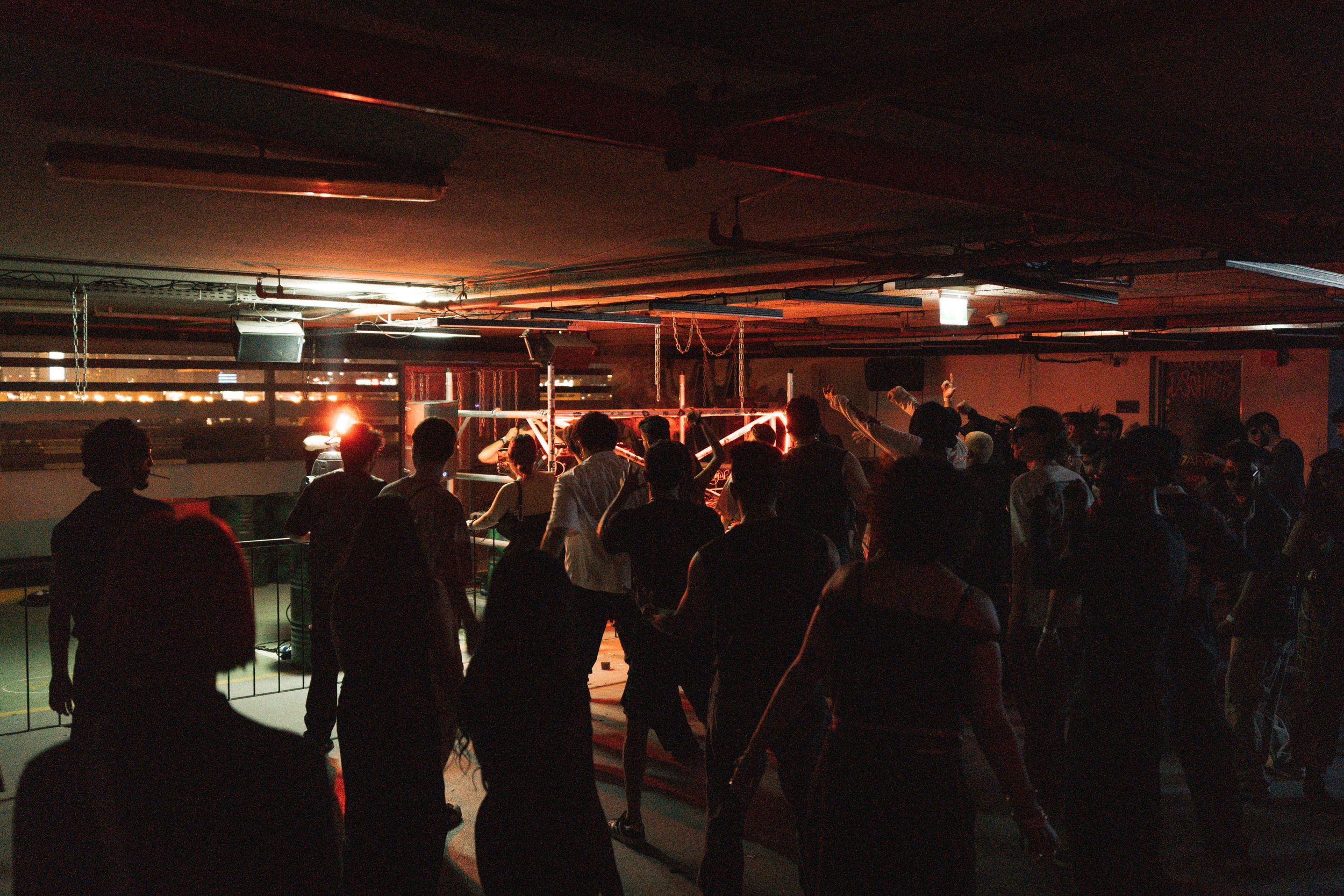 A group of people dancing and socializing at a dimly lit indoor party or nightclub with red neon lighting.