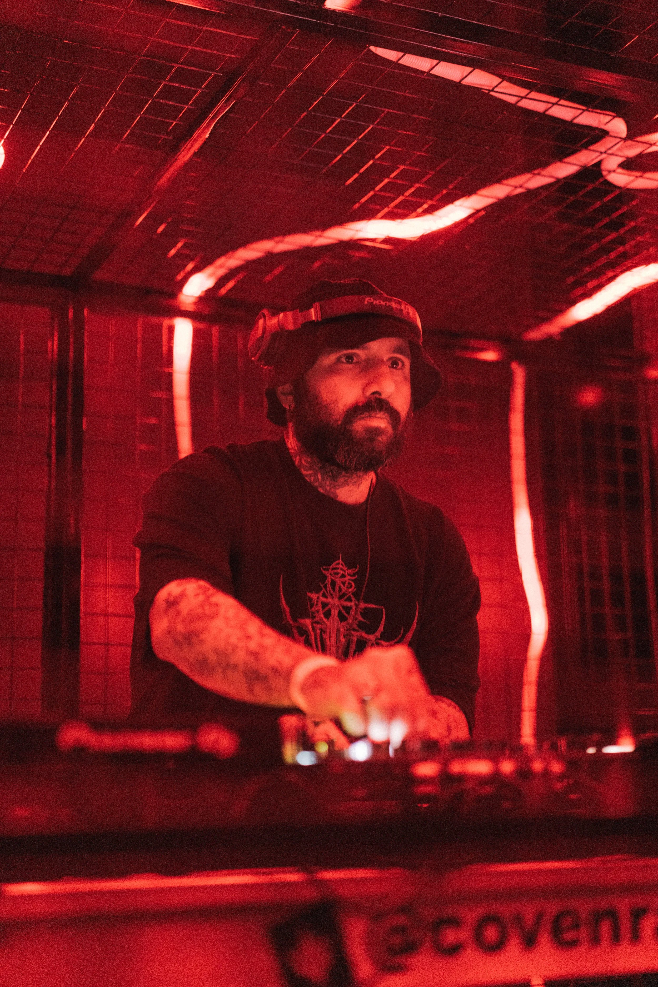 A DJ with a beard and tattoos wearing headphones performs in a dimly lit space with red lighting.