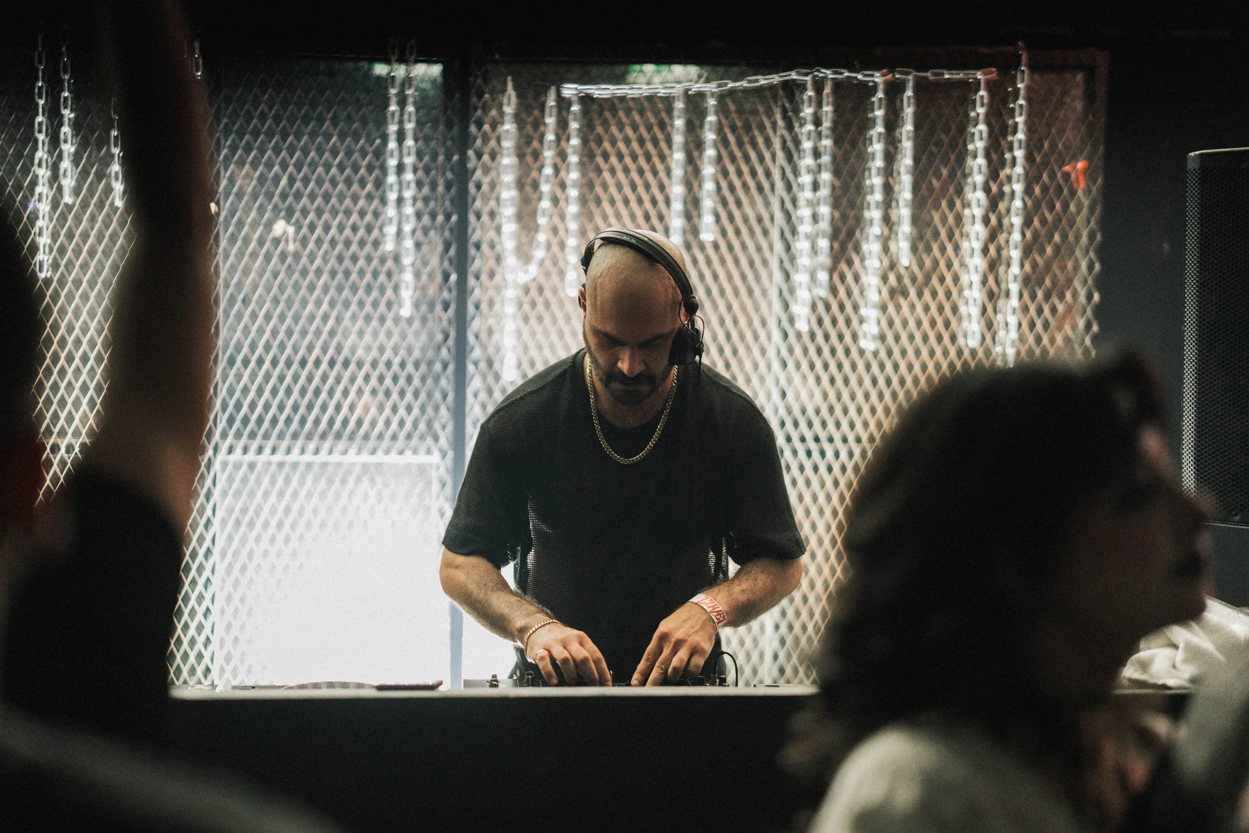 A DJ with a shaved head, wearing headphones, a black shirt, and a gold chain, is mixing music at a nightclub or event, with a patterned backdrop and dim lighting.