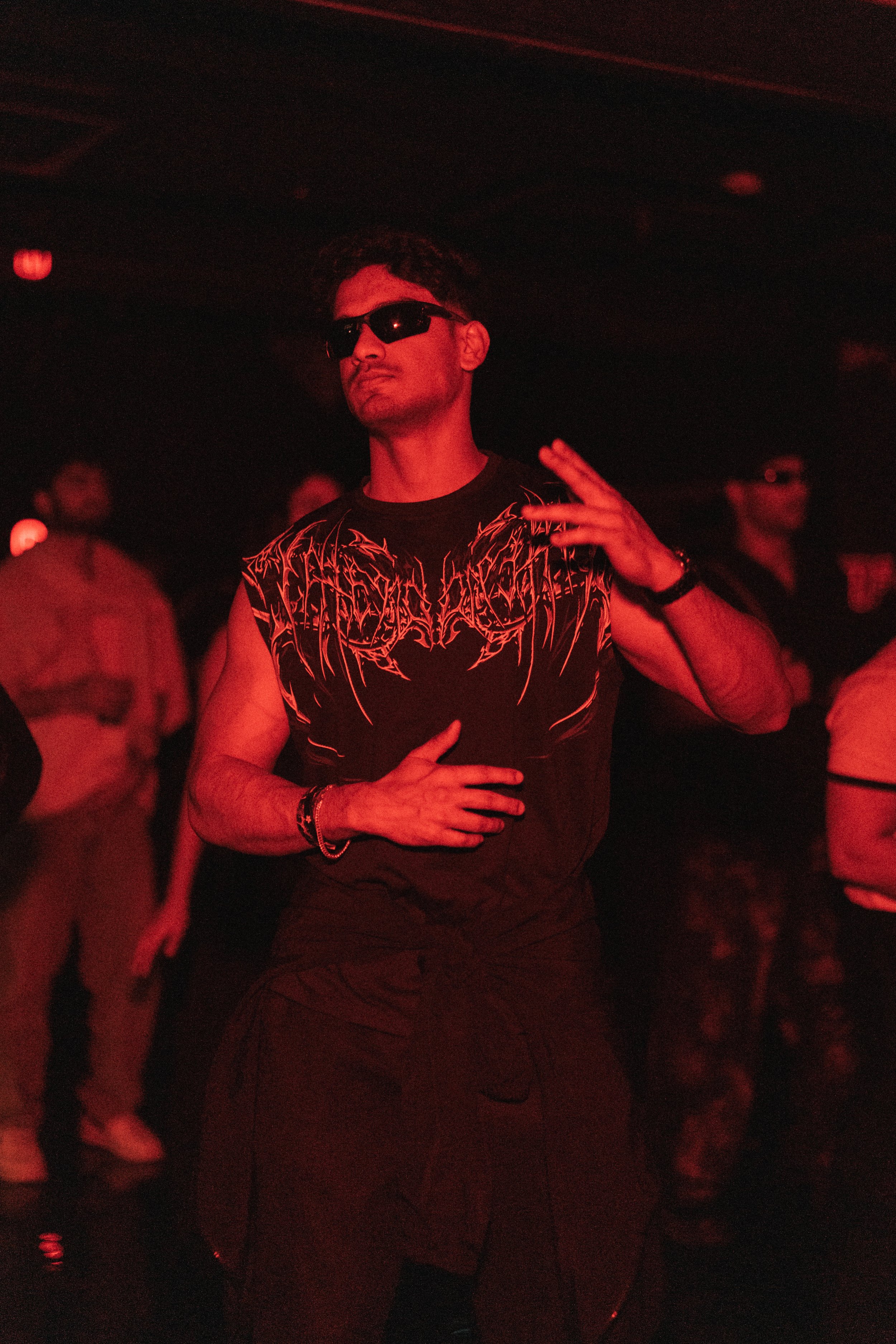 A person dancing at a concert or club, wearing sunglasses, a sleeveless shirt, and dark pants, with other people in the background.