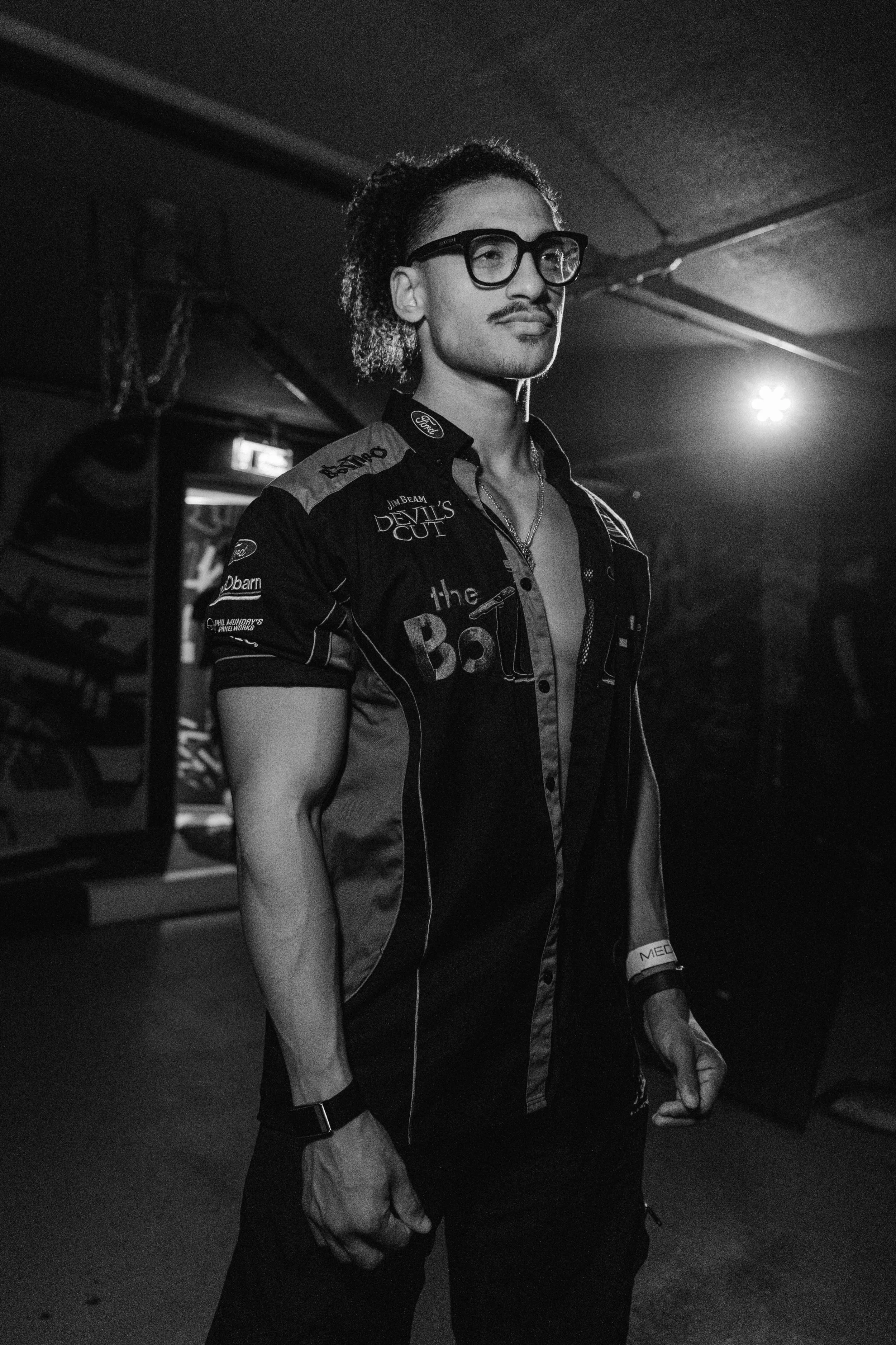 A young man with glasses and a mustache wearing a racing jacket with sponsor logos, standing in a dimly lit room.