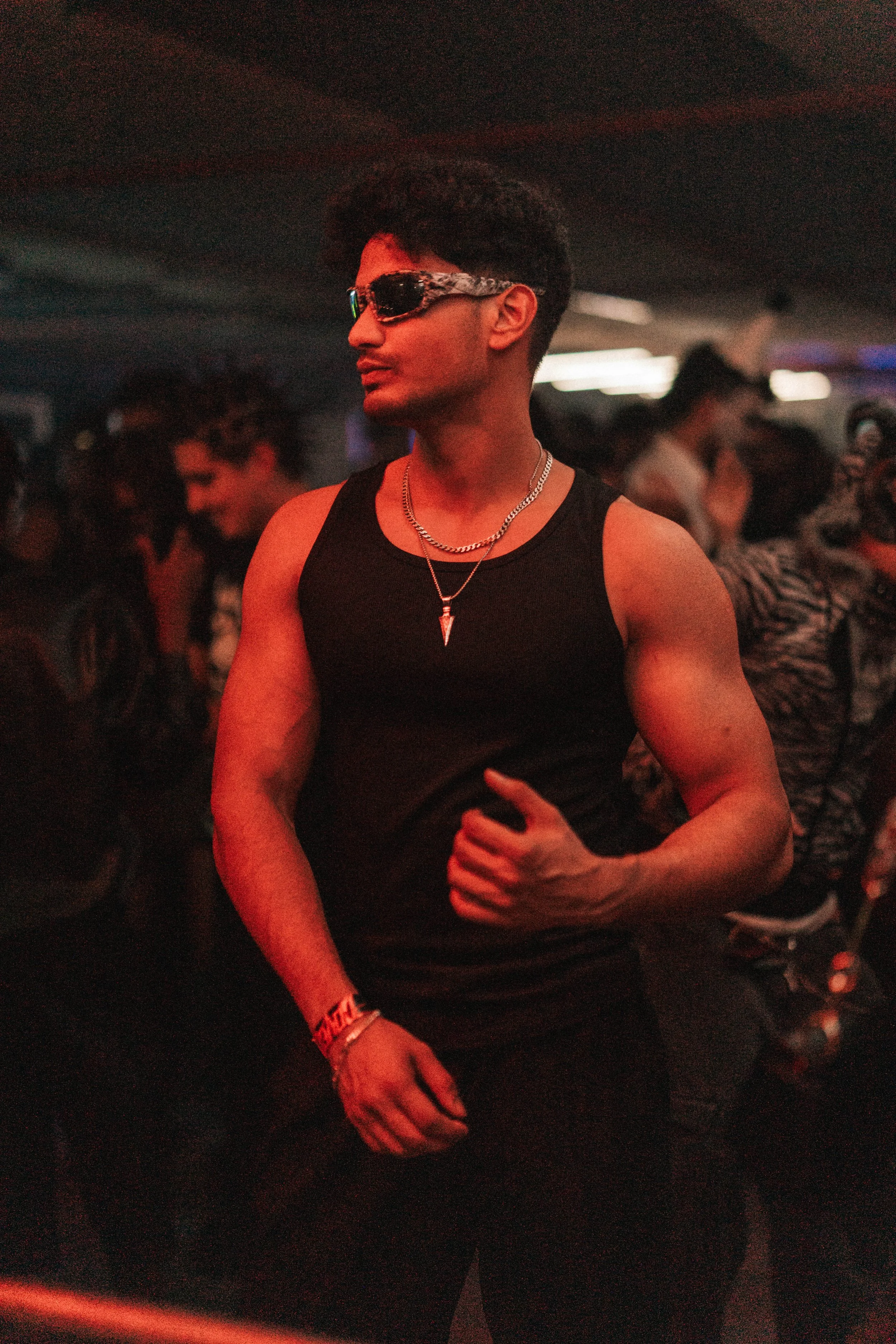 A young man with dark hair wearing sunglasses, a black sleeveless shirt, silver necklaces, and a wristband, standing in a dimly lit party or club with other people in the background.