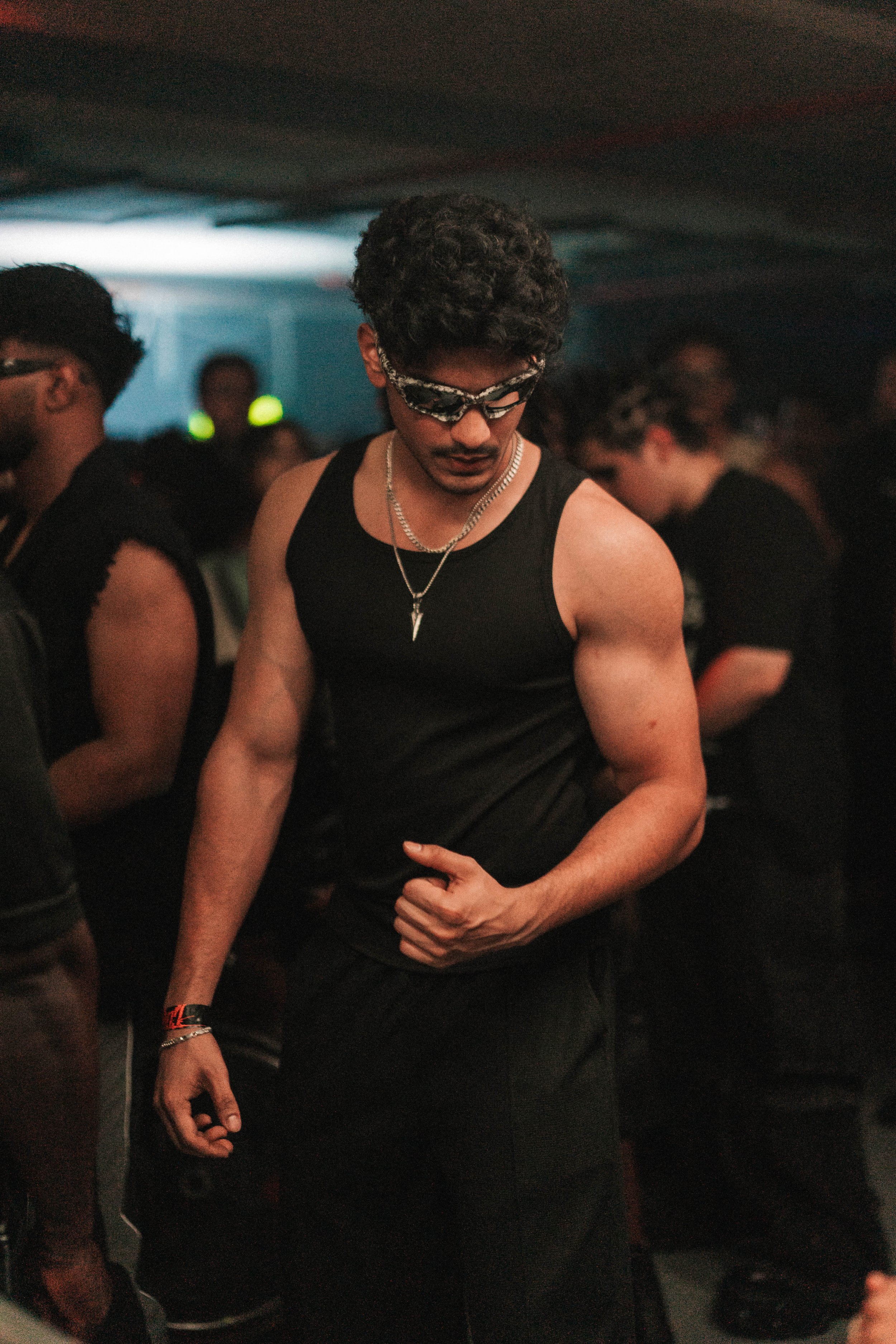 A man wearing sunglasses, a black sleeveless shirt, and silver necklaces is dancing at a dark party or club, surrounded by other people.