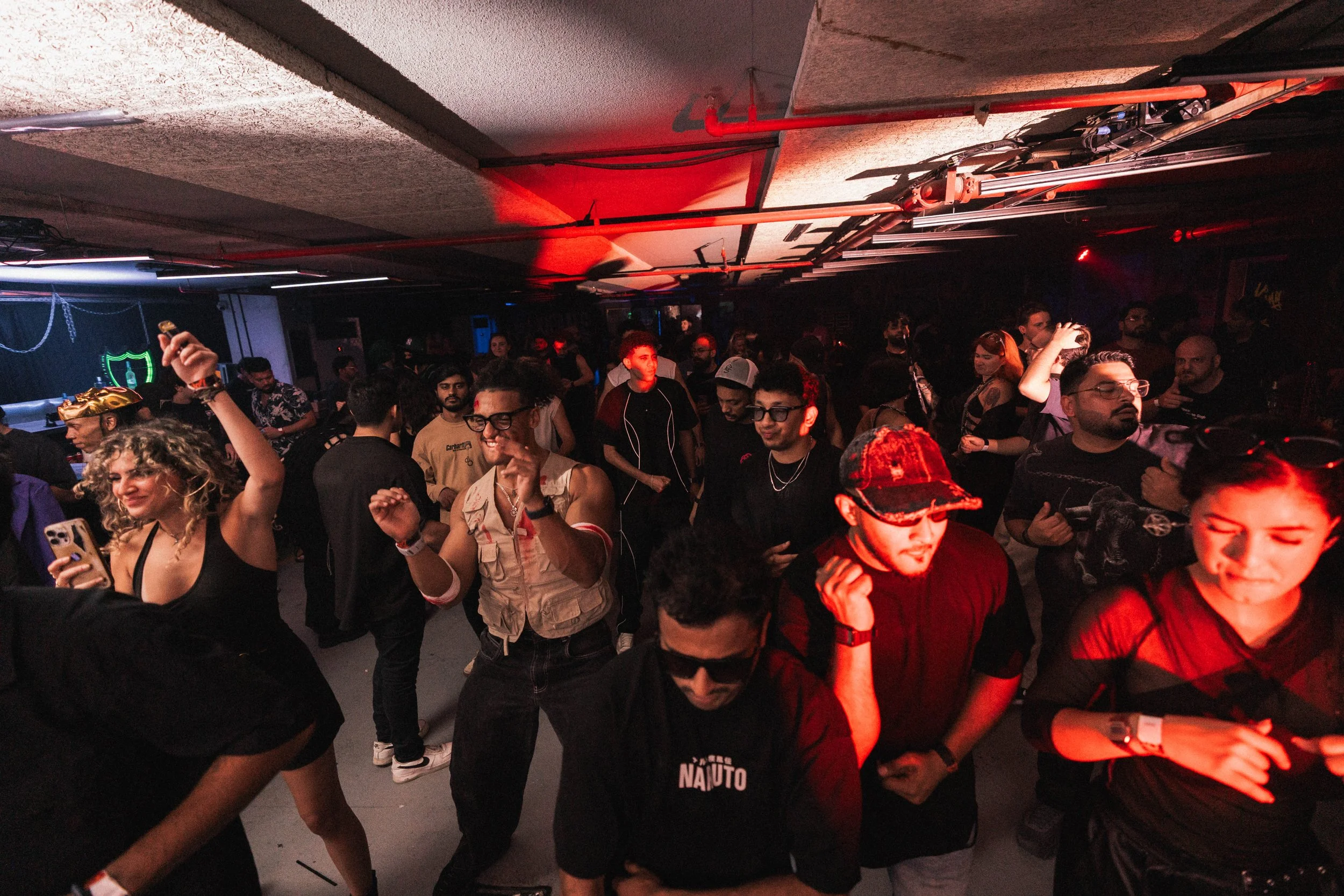 People dancing and enjoying music at a crowded nightclub with red lighting and a DJ booth in the background.