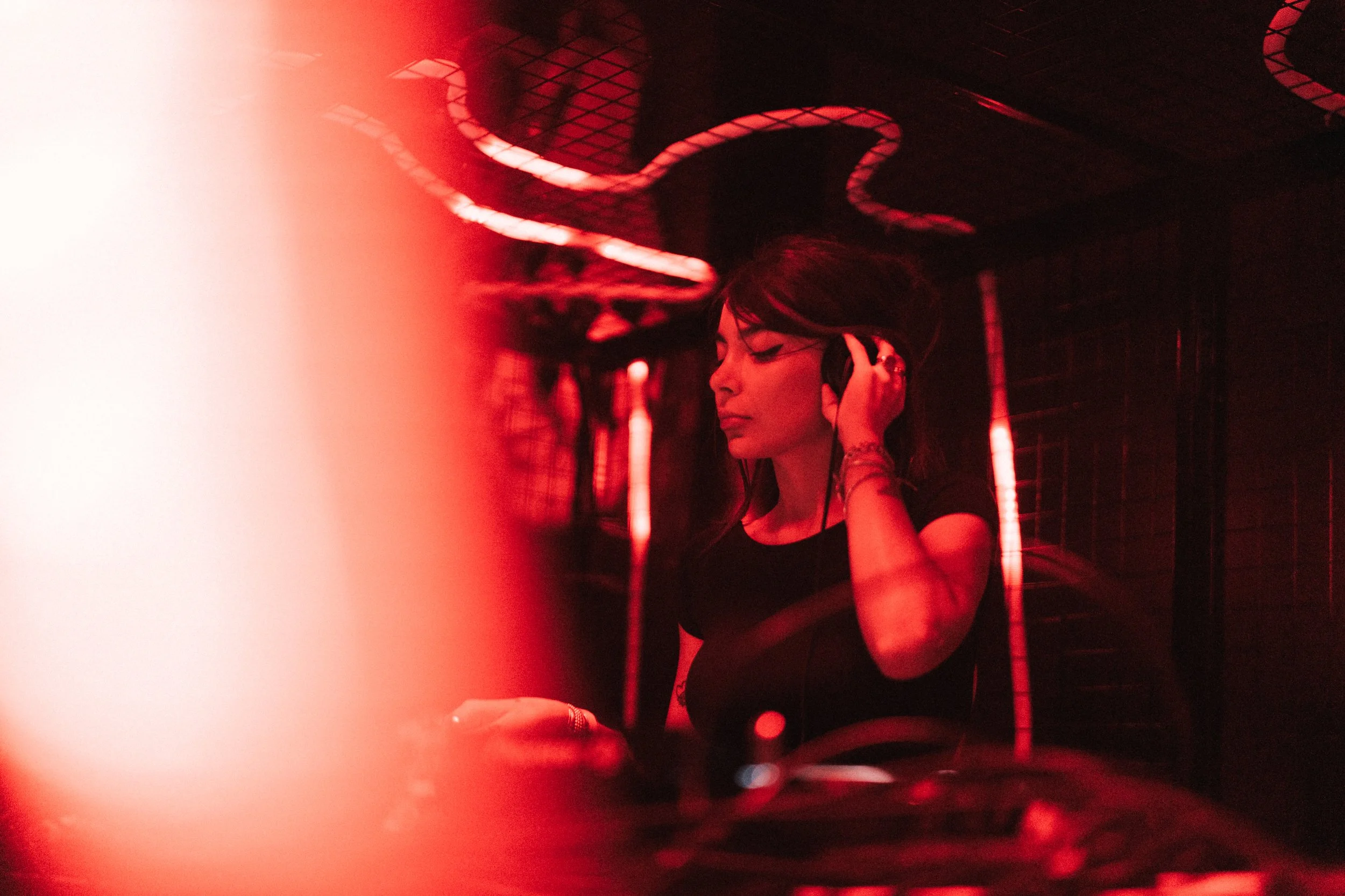 A woman wearing headphones, with long dark hair and makeup, in a dark room illuminated by red neon lights, possibly a nightclub or music venue.