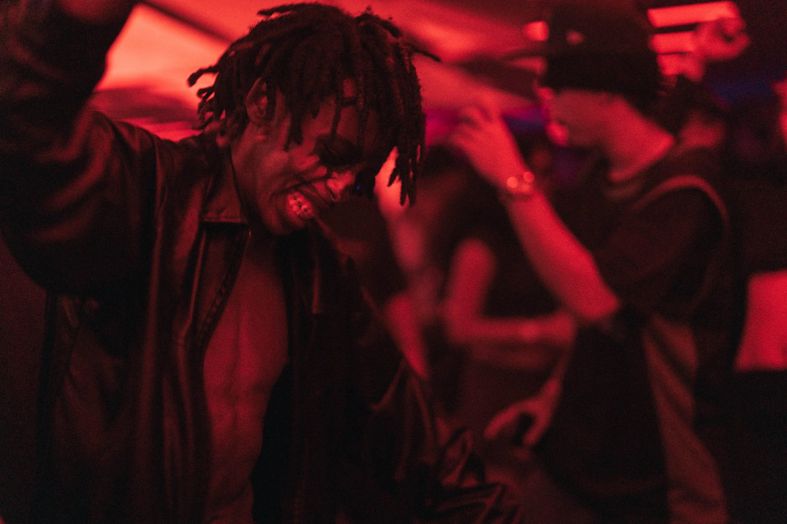 A person dancing in a dimly lit environment with red lighting, wearing glasses and a leather jacket, smiling and enjoying the moment, surrounded by other people.