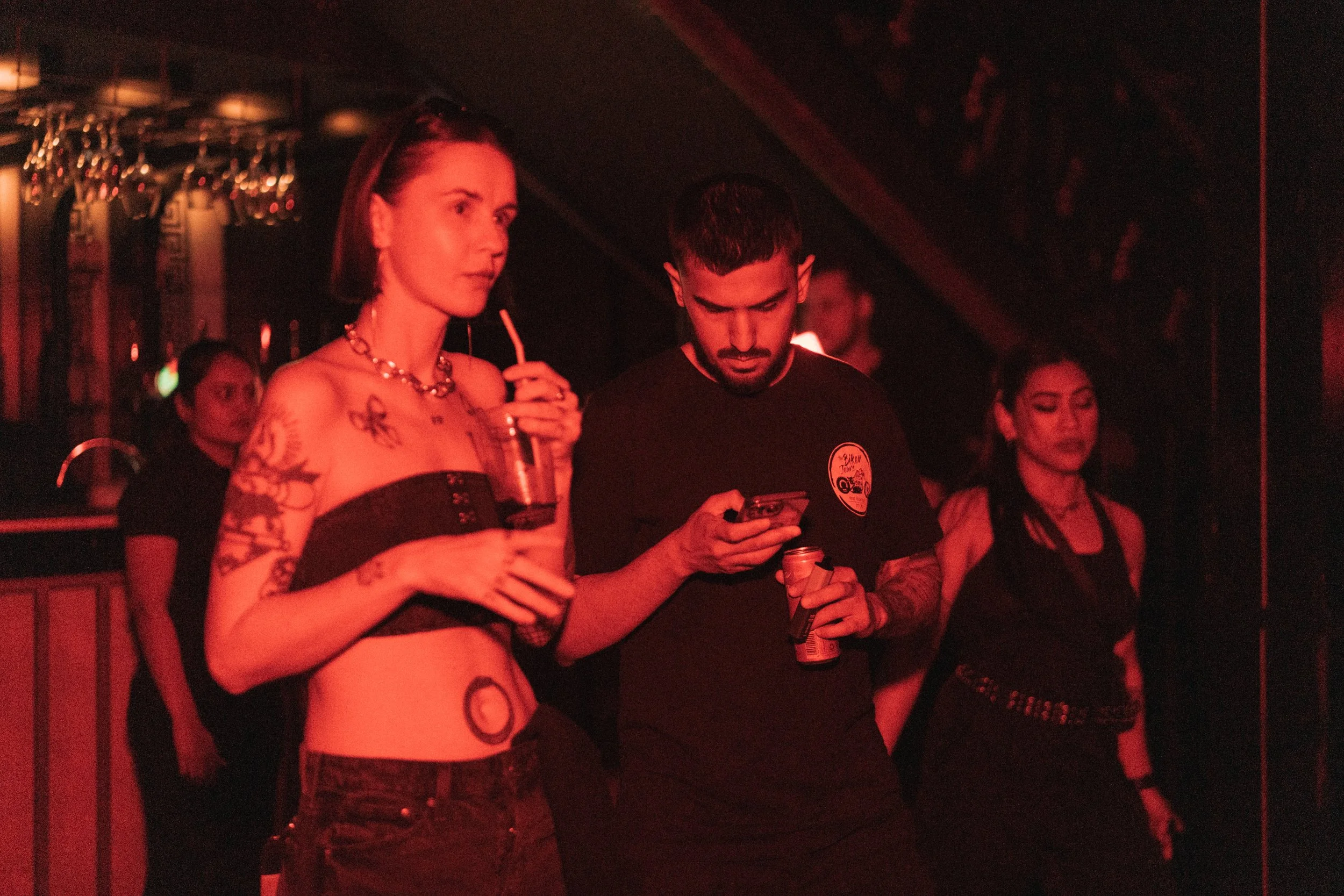 A woman with tattoos and short hair holding a drink, next to a man looking at his phone, in a dark, red-lit bar or club environment.