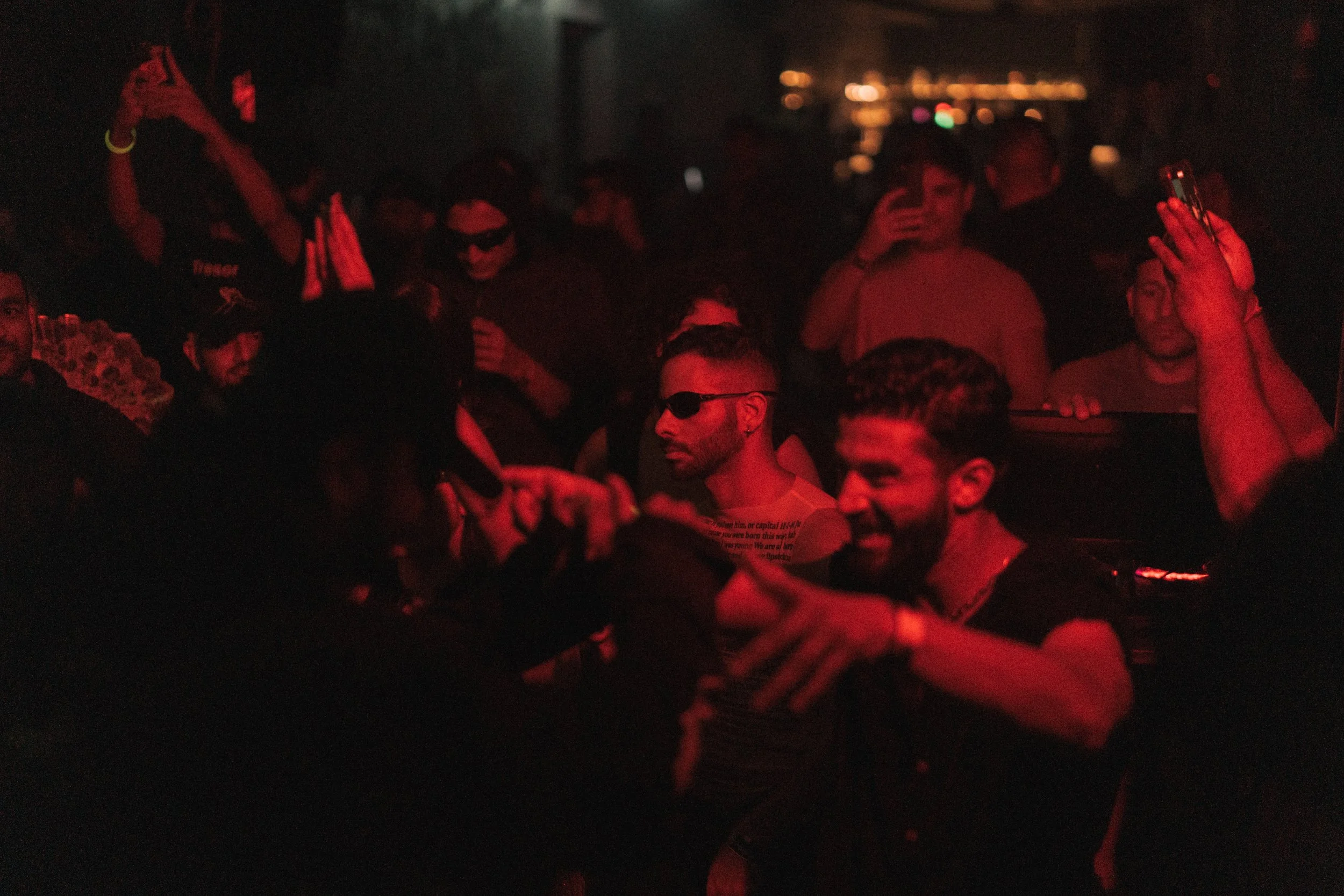 People at a nightclub or concert, illuminated by red lighting, some taking photos or videos, others dancing or socializing.