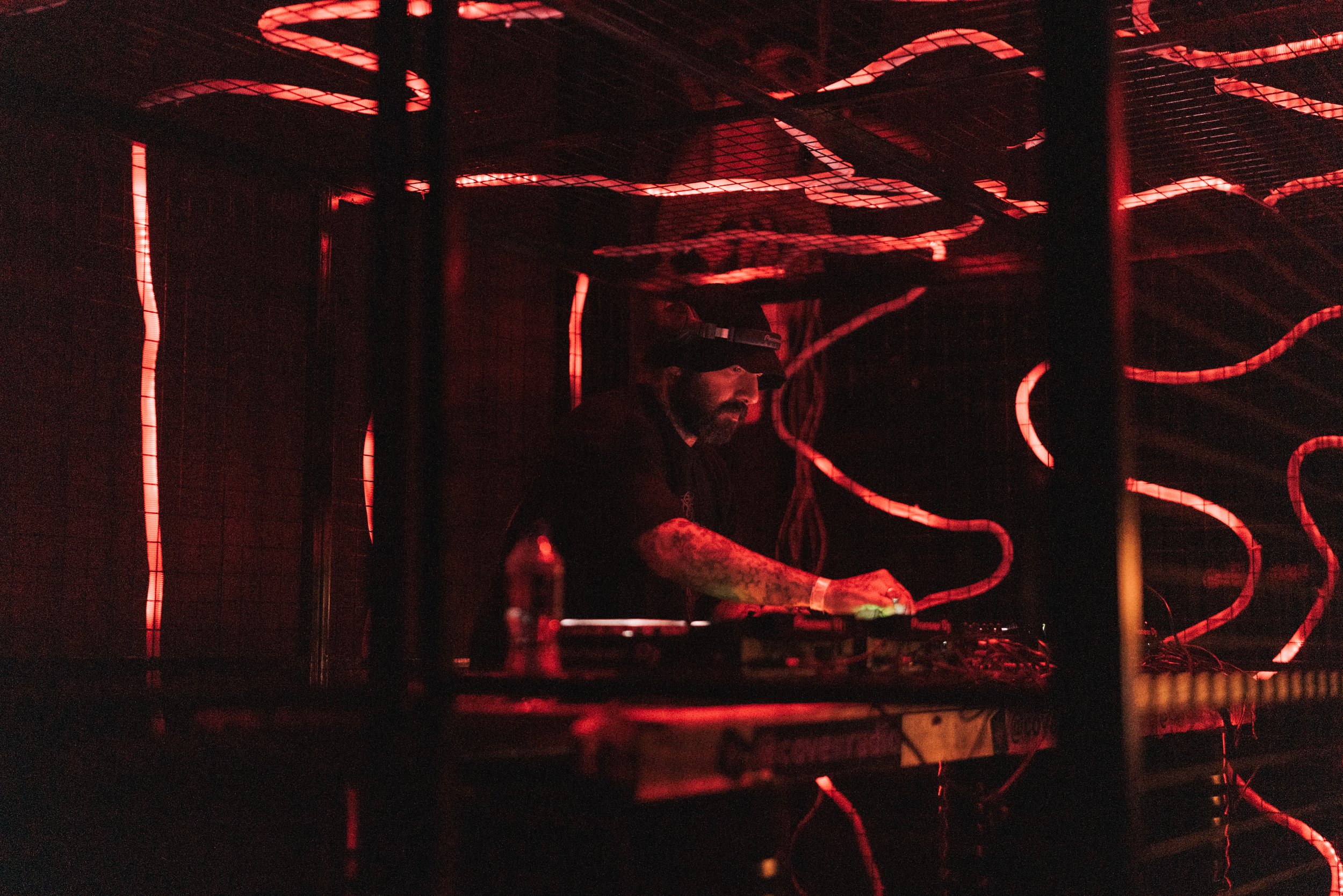 A DJ performing in a dark, red-lit space with glowing red and pink neon lights on the ceiling, behind a metal grid.