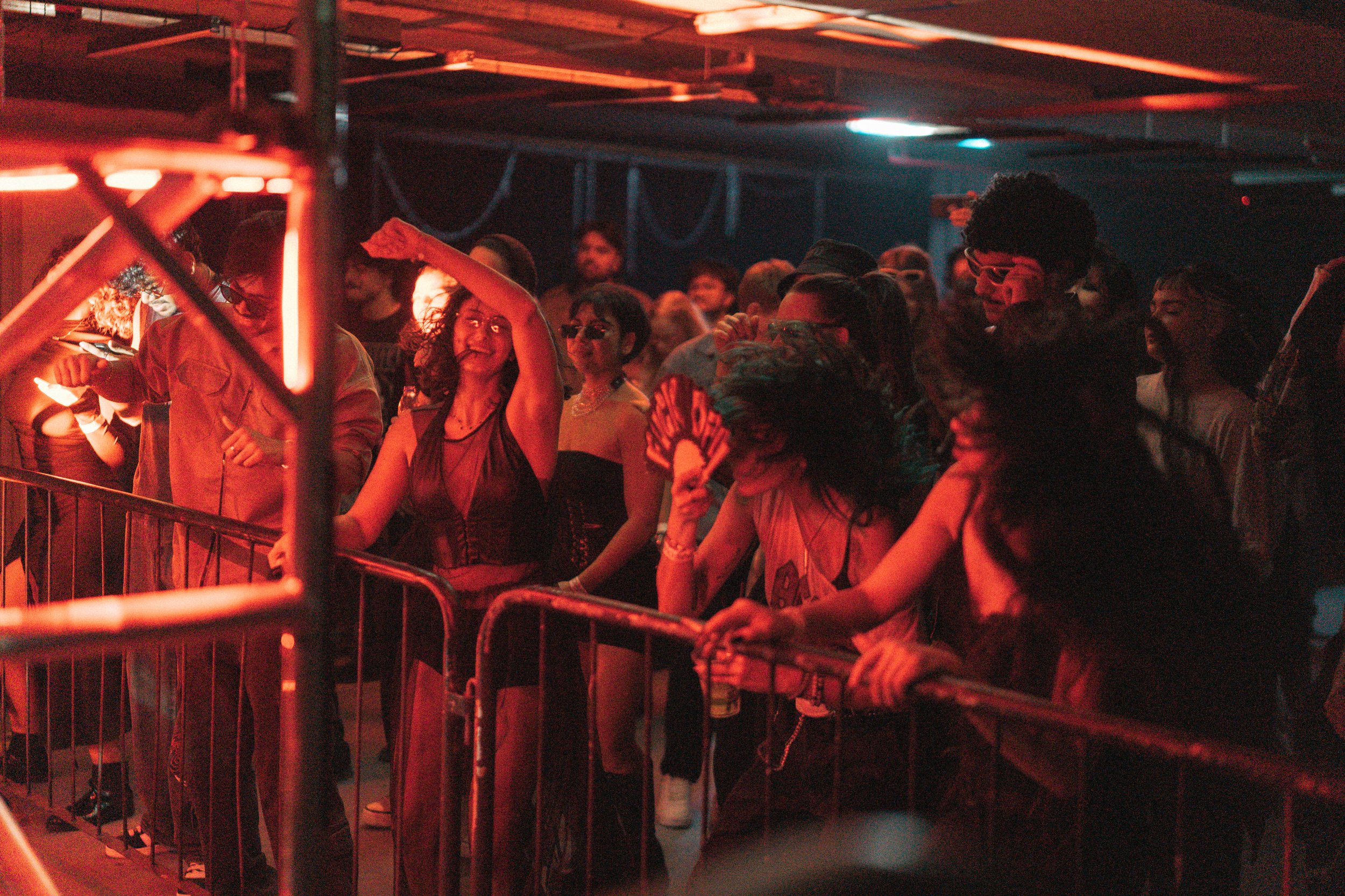 People dancing and enjoying music at a nightclub with red lighting and a metal railing.