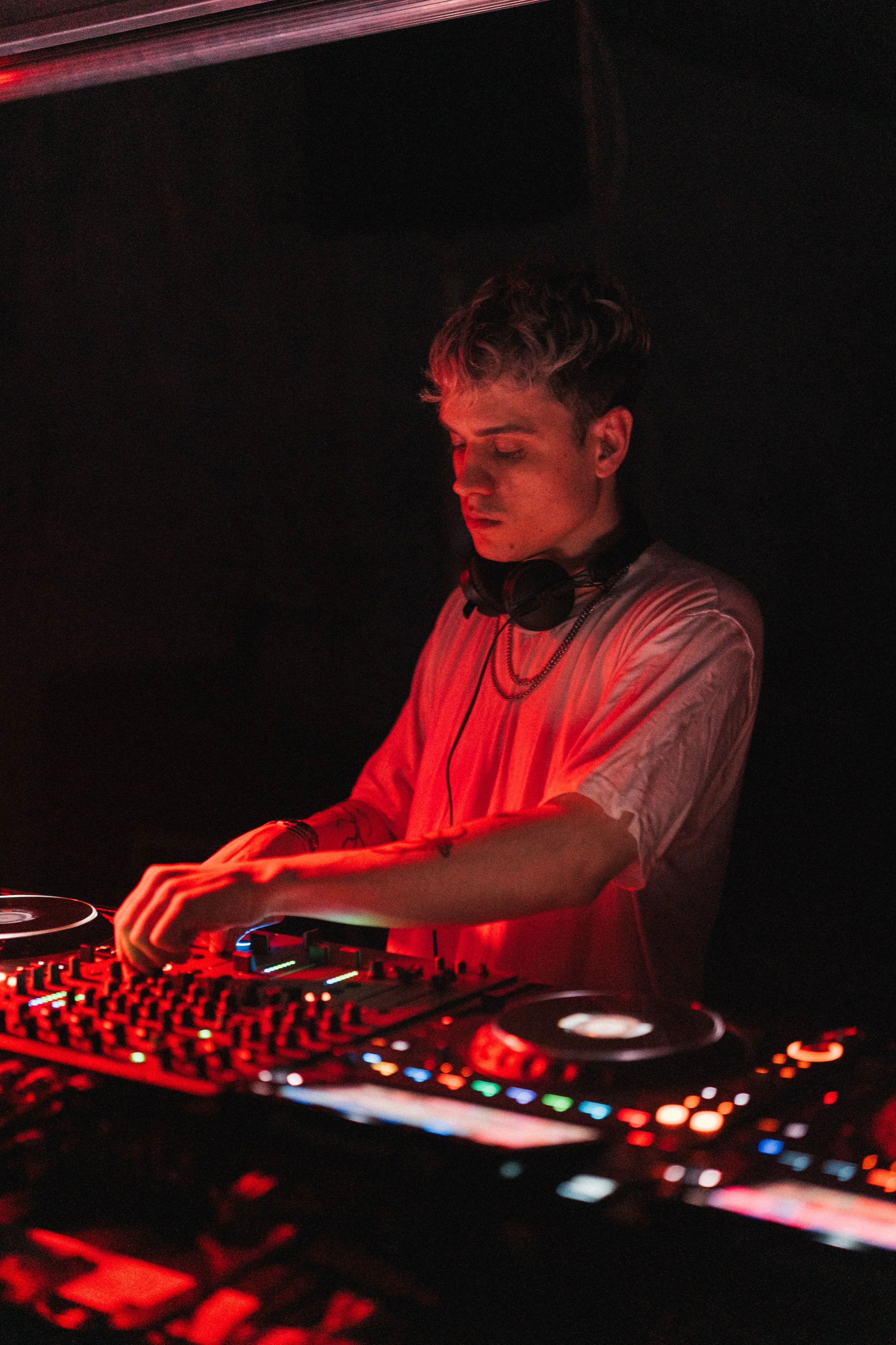 A young male DJ with headphones around his neck mixing music at a DJ booth in a dark club, illuminated by red lighting.