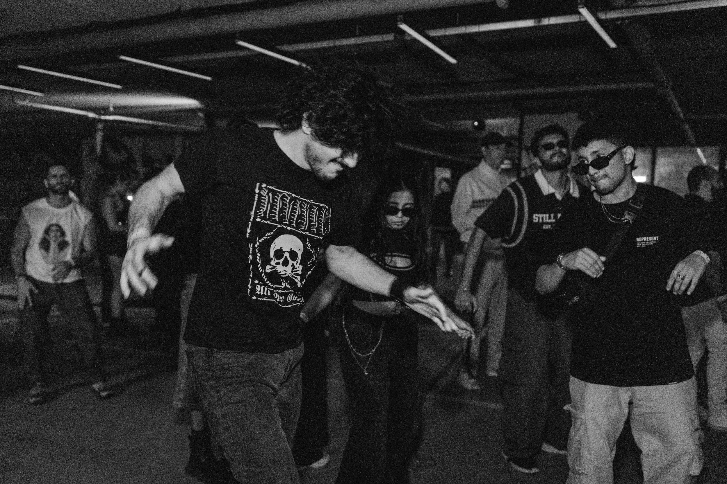 People dancing in a dark indoor venue, some wearing sunglasses, with an energetic atmosphere.