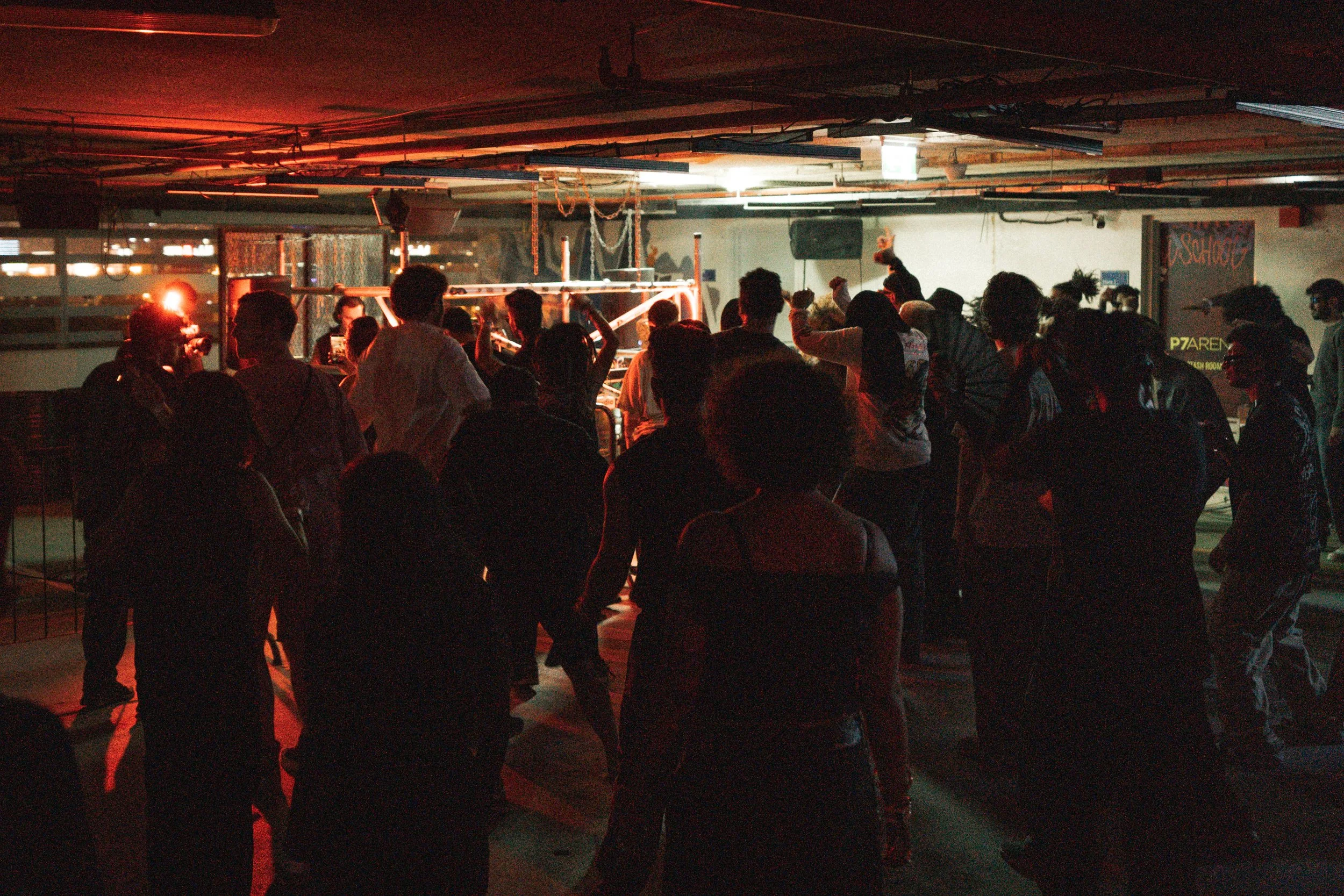 People dancing and socializing in a dimly lit indoor space with a DJ playing music, illuminated by red and warm lighting.