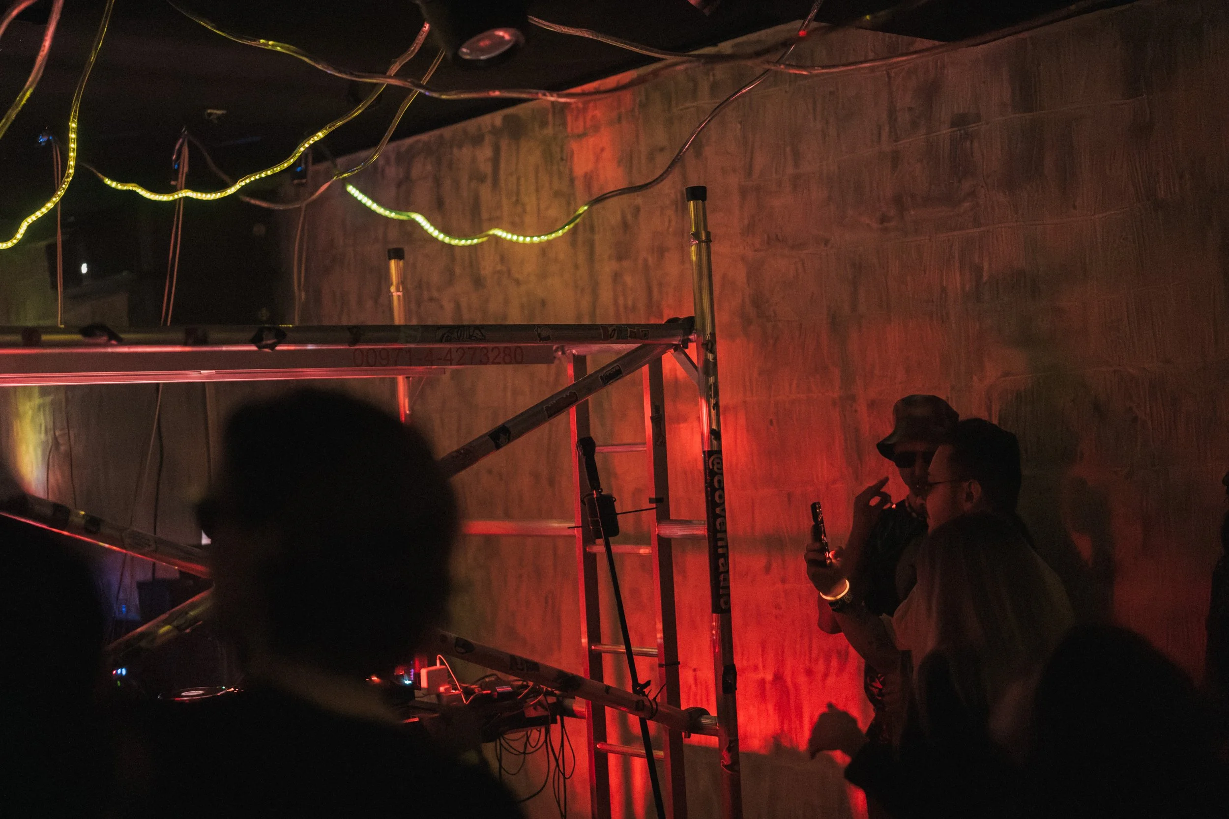 Two people with sunglasses and a hat enjoying drinks at a dark, indoor party with red and green LED lights, ladder, and scattered silhouettes of other people.