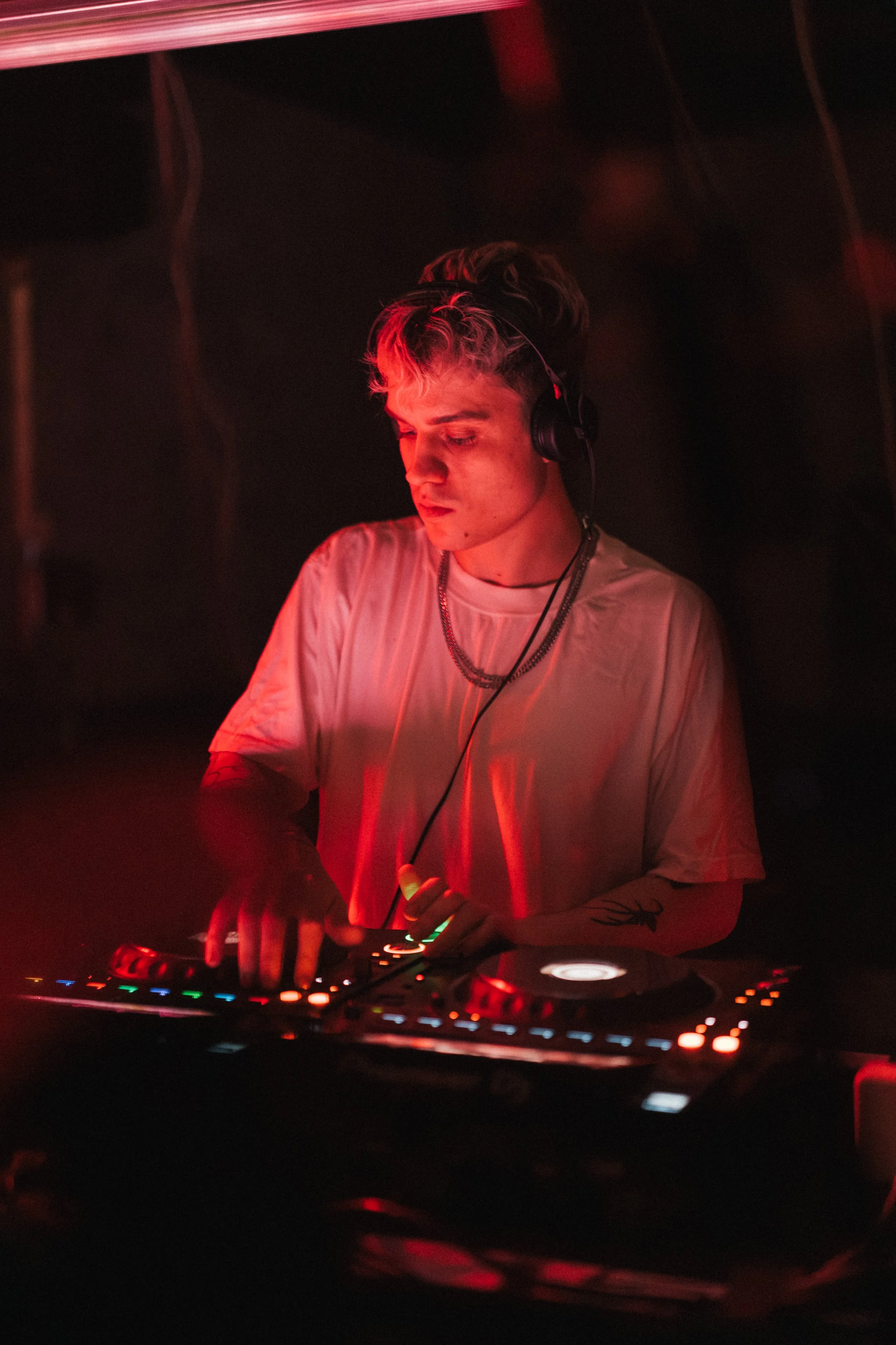Young male DJ performing with illuminated DJ equipment in a dimly lit setting, wearing headphones and a white t-shirt.