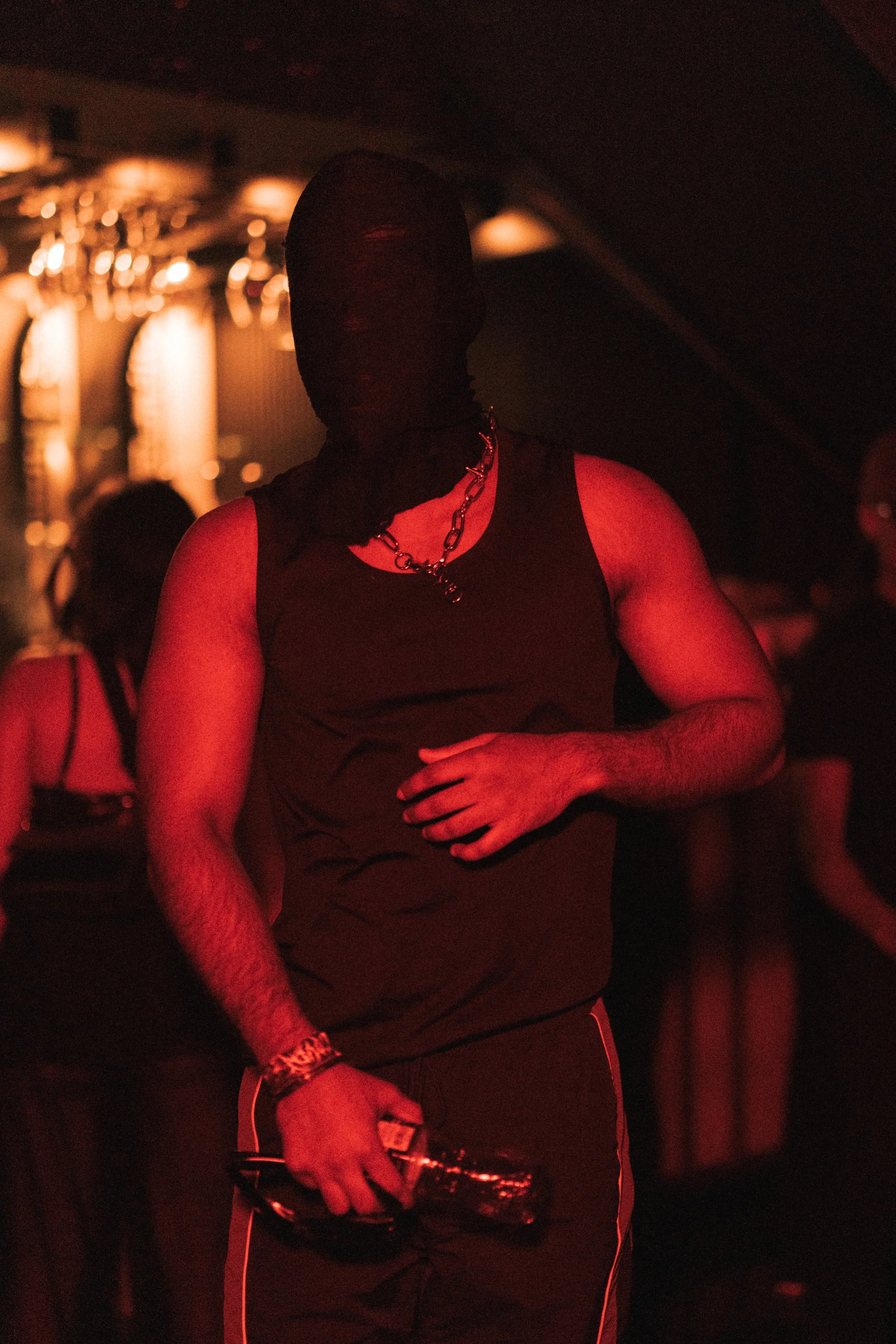 Person wearing a black ski mask, sleeveless black shirt, holding a glass bottle, in a dimly lit club with red lighting and blurred background of other people.