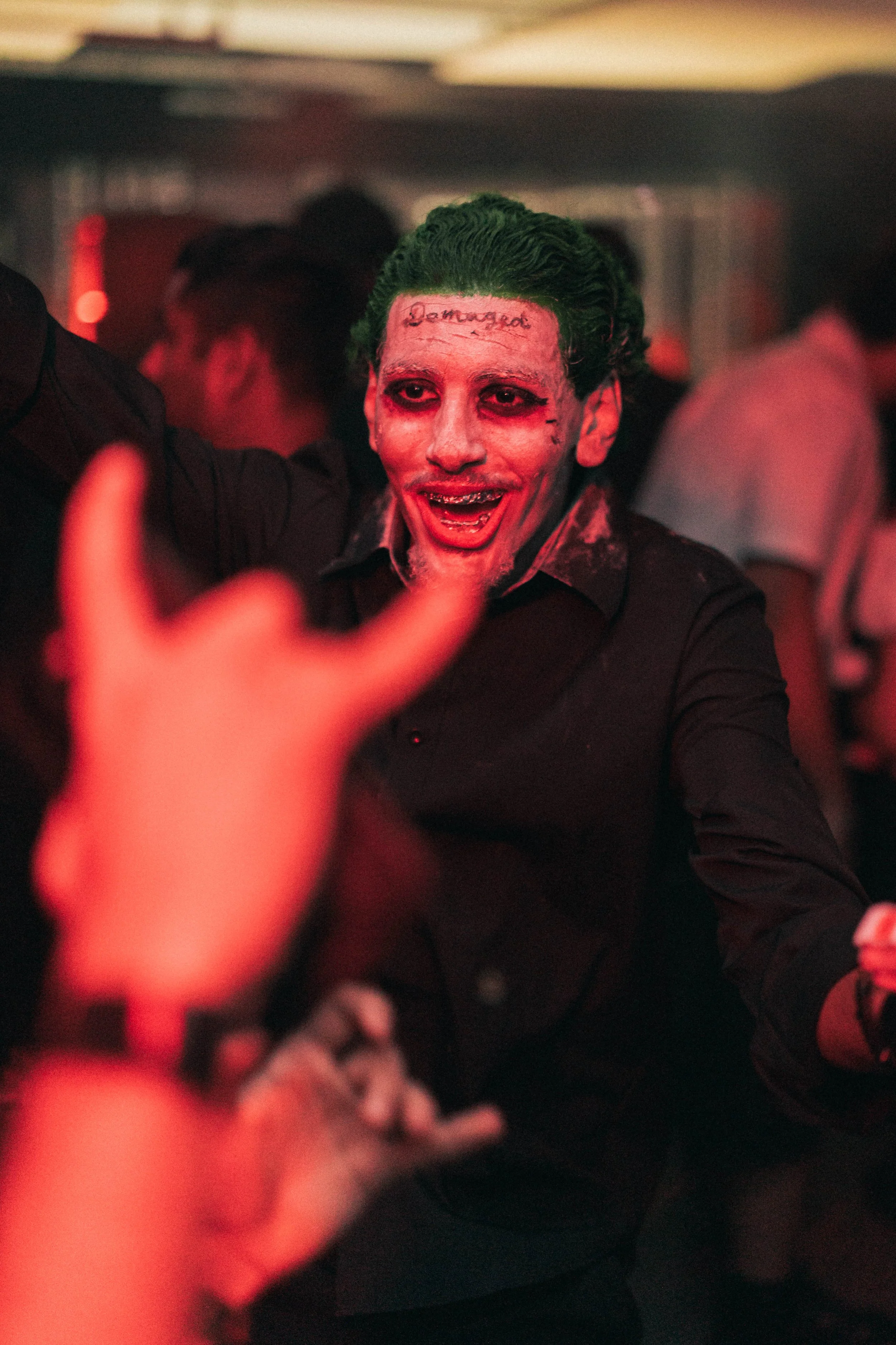 A man with green hair and face makeup resembles the Joker character, smiling and reaching out with his hands while dancing at a party or club with red lighting and other people in the background.