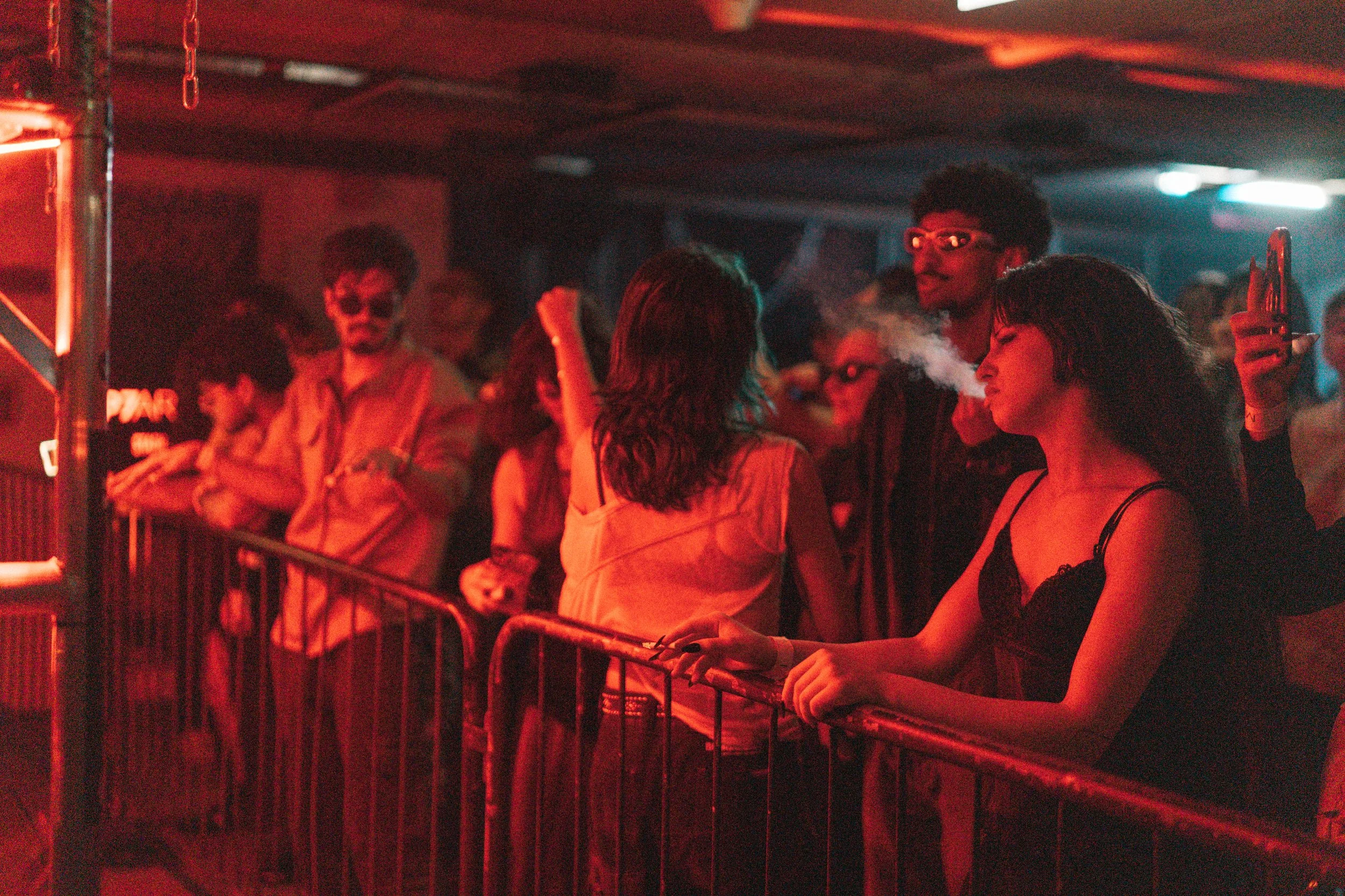 People standing behind a metal barrier at a nightclub, illuminated by red and orange lights, some smoking and chatting.
