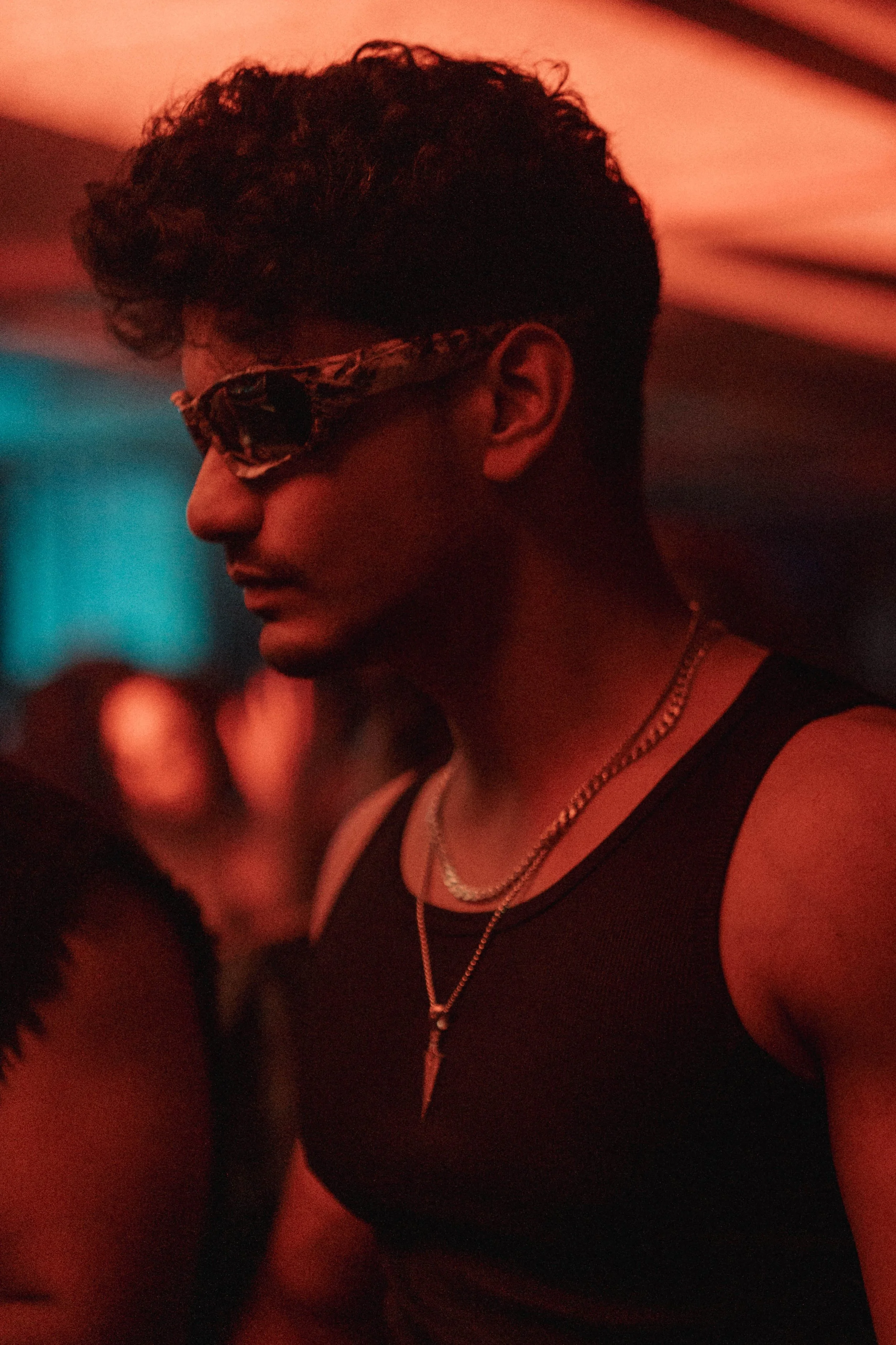 A person with dark curly hair wearing sunglasses with animal print, a black tank top, and layered necklaces, in a dimly lit setting with red and blue lighting.