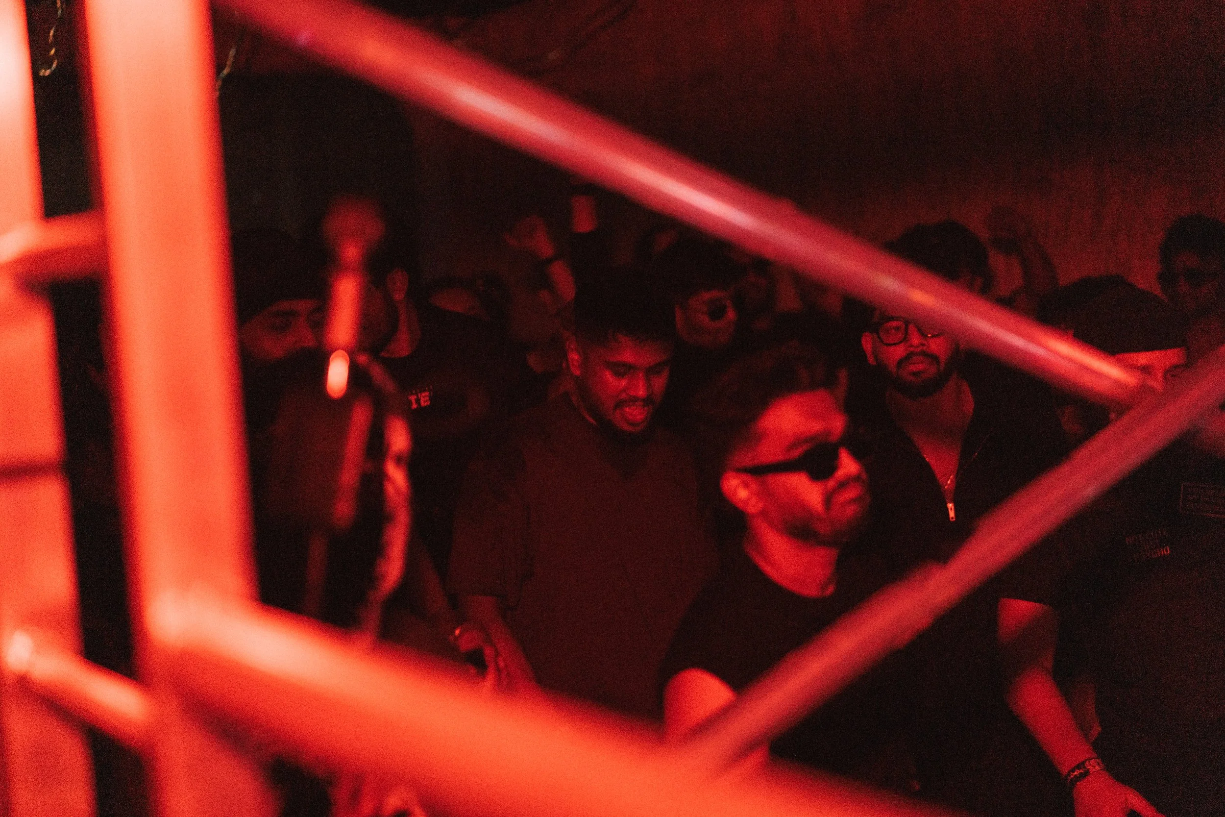 People at a concert or nightclub illuminated by red lighting, shown through metal bars in the foreground.