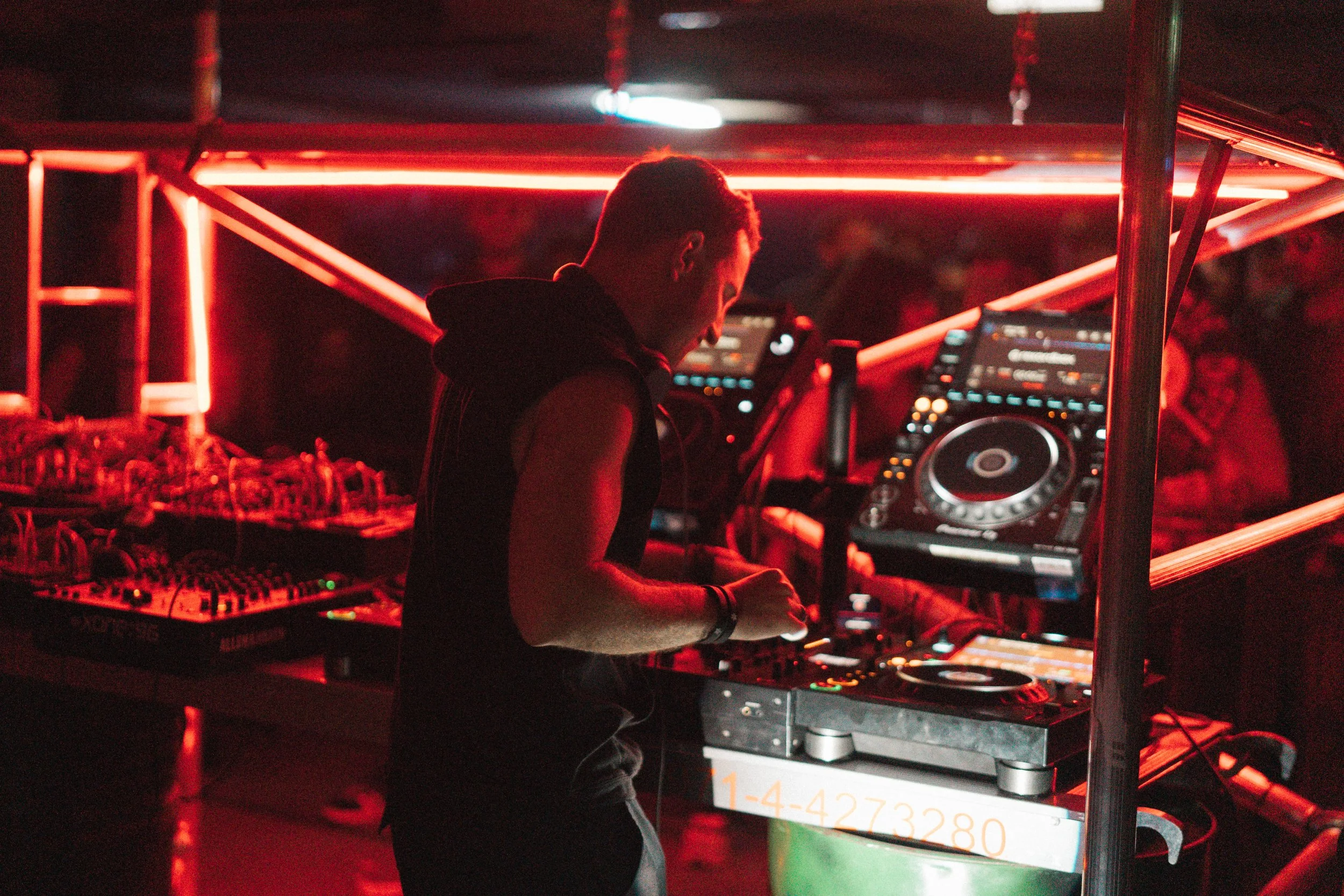 A DJ performing at a nightclub, illuminated by red lighting, using turntables and mixers behind a DJ booth.