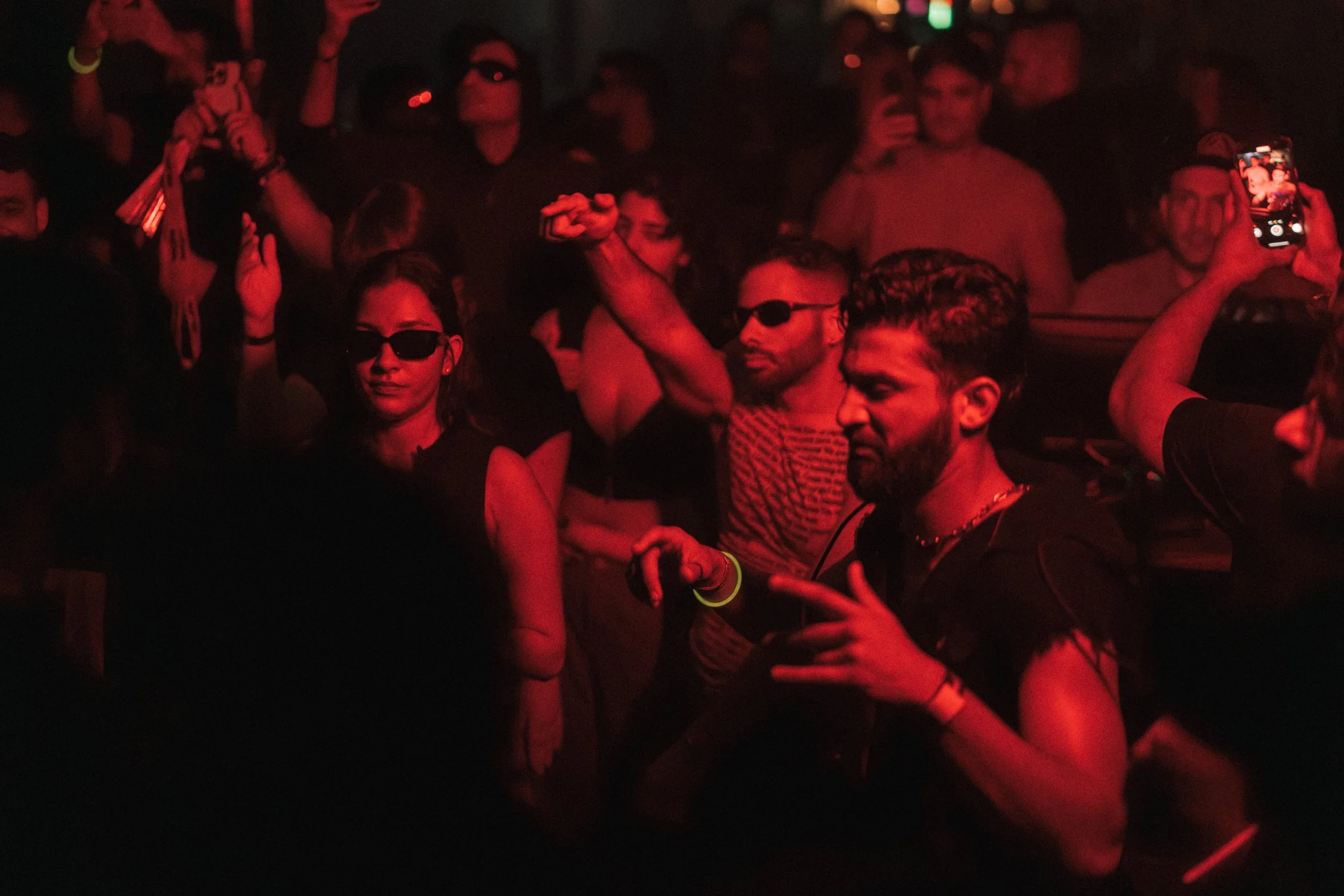 People dancing and enjoying themselves at a nightclub with red lighting, some wearing sunglasses, many with drinks, and a person taking a photo with their phone.