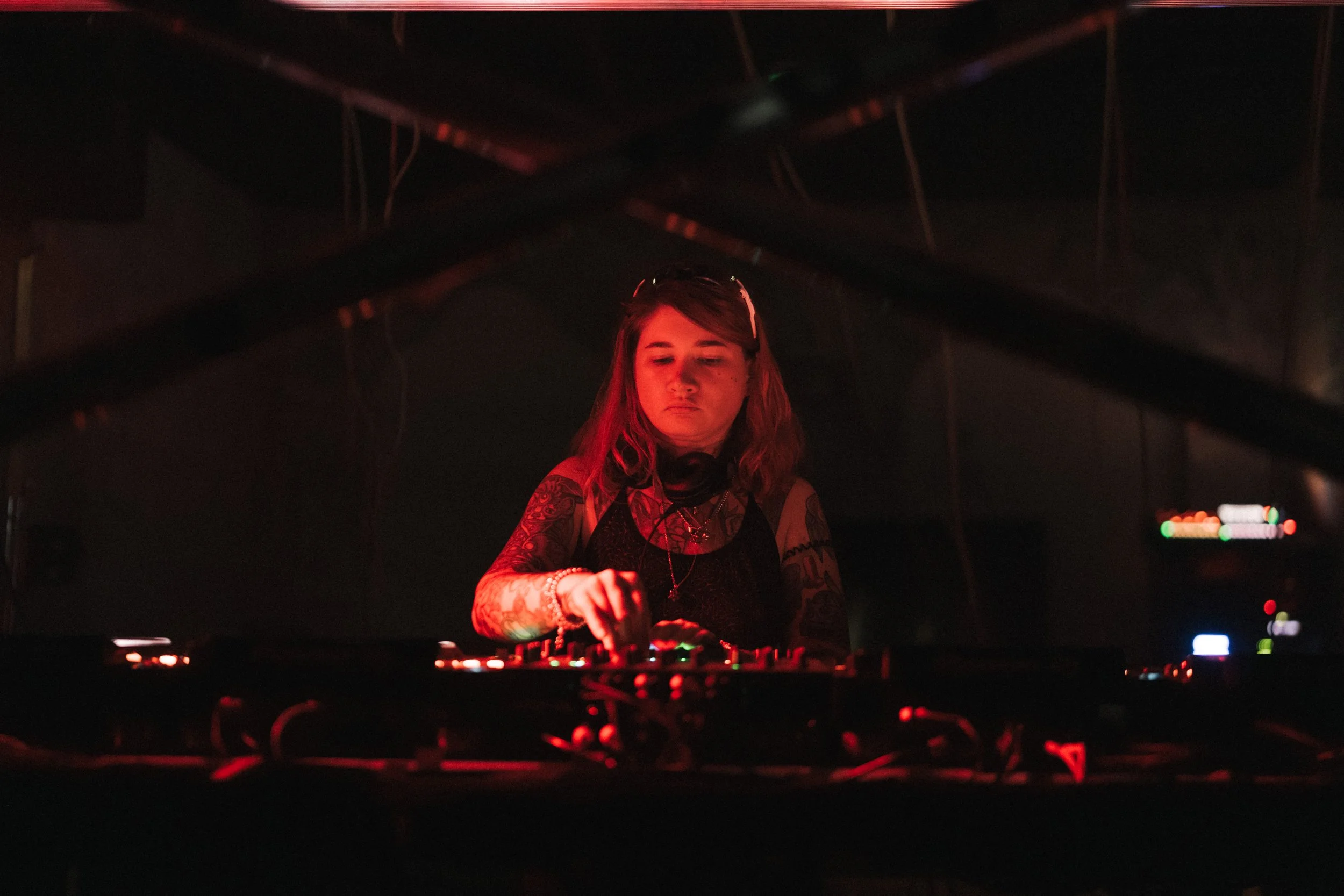 A female DJ with tattoos on her arms wearing headphones around her neck, performing at a dimly lit event with red lighting and equipment in front of her.