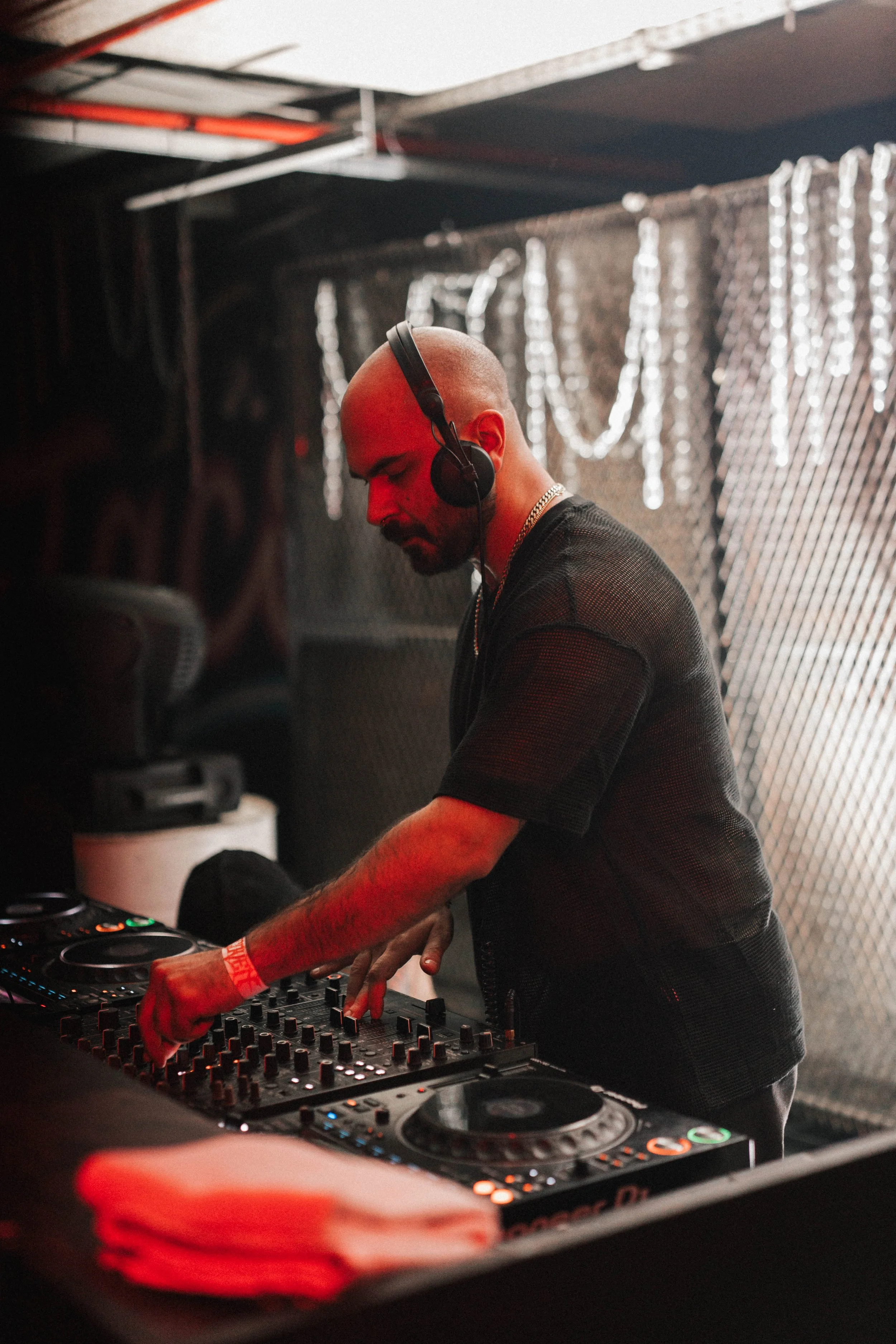 A male DJ wearing headphones and a black shirt, mixing music on a DJ controller at a party or club, with dim lighting and decorative backdrop.