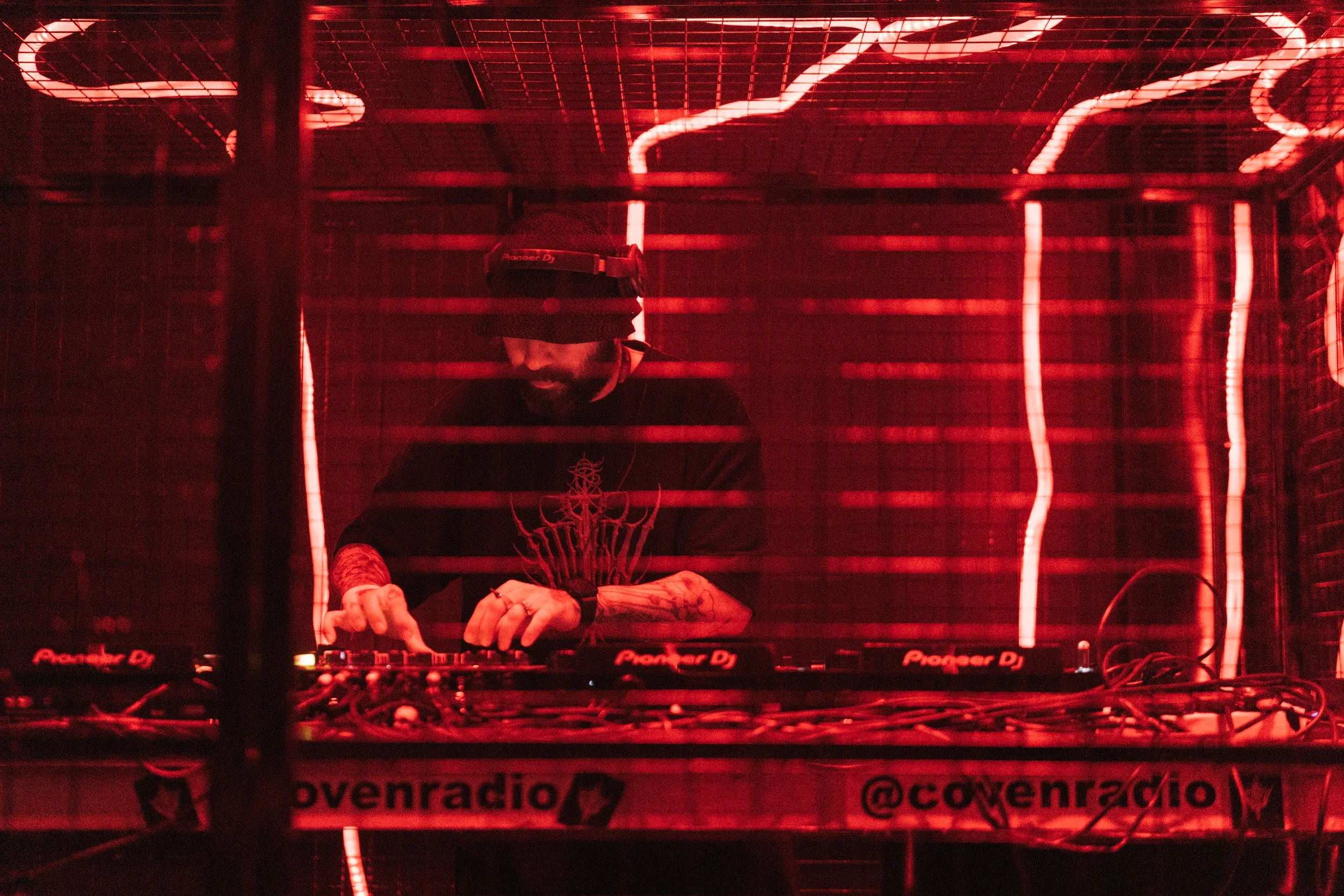 DJ performing behind red lighting with neon light art on the wall, wearing a black shirt with a graphic design, using DJ equipment in a dark room.