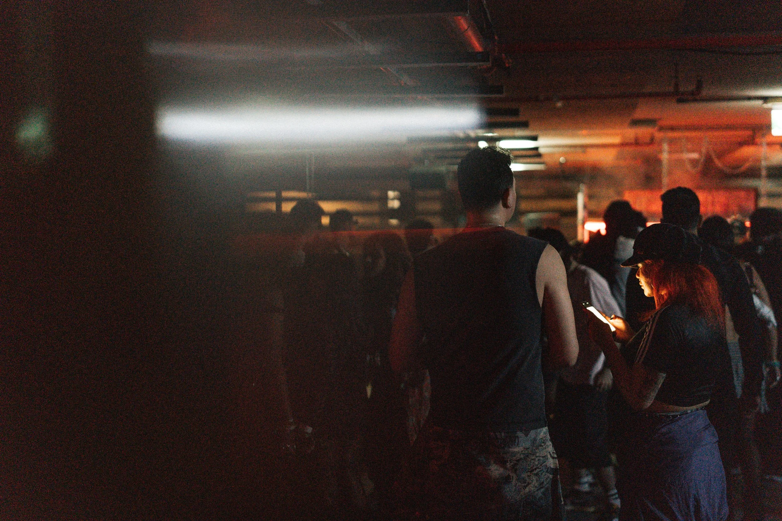 A dimly lit indoor space with people waiting or standing, illuminated by soft red and white lighting. A person on their phone is visible, and most of the crowd is facing away from the camera.